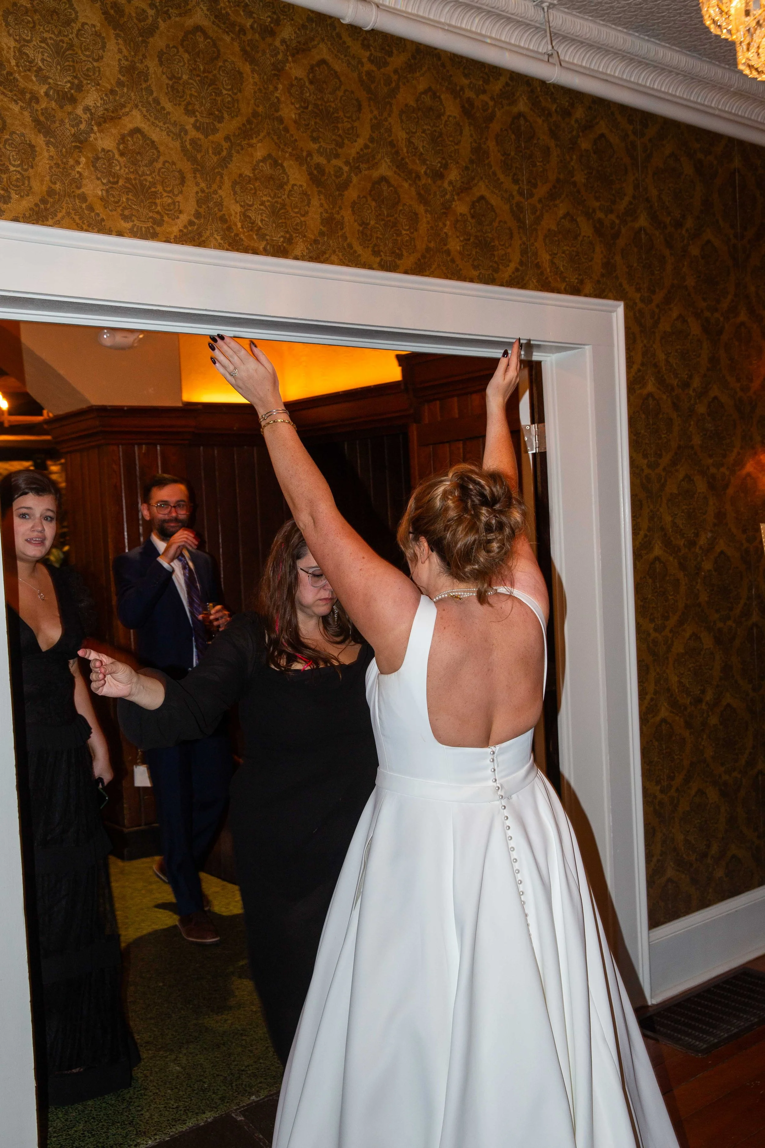 A woman in a white wedding dress dances with arms raised in front of a mirror at a wedding reception, with guests watching in the background.