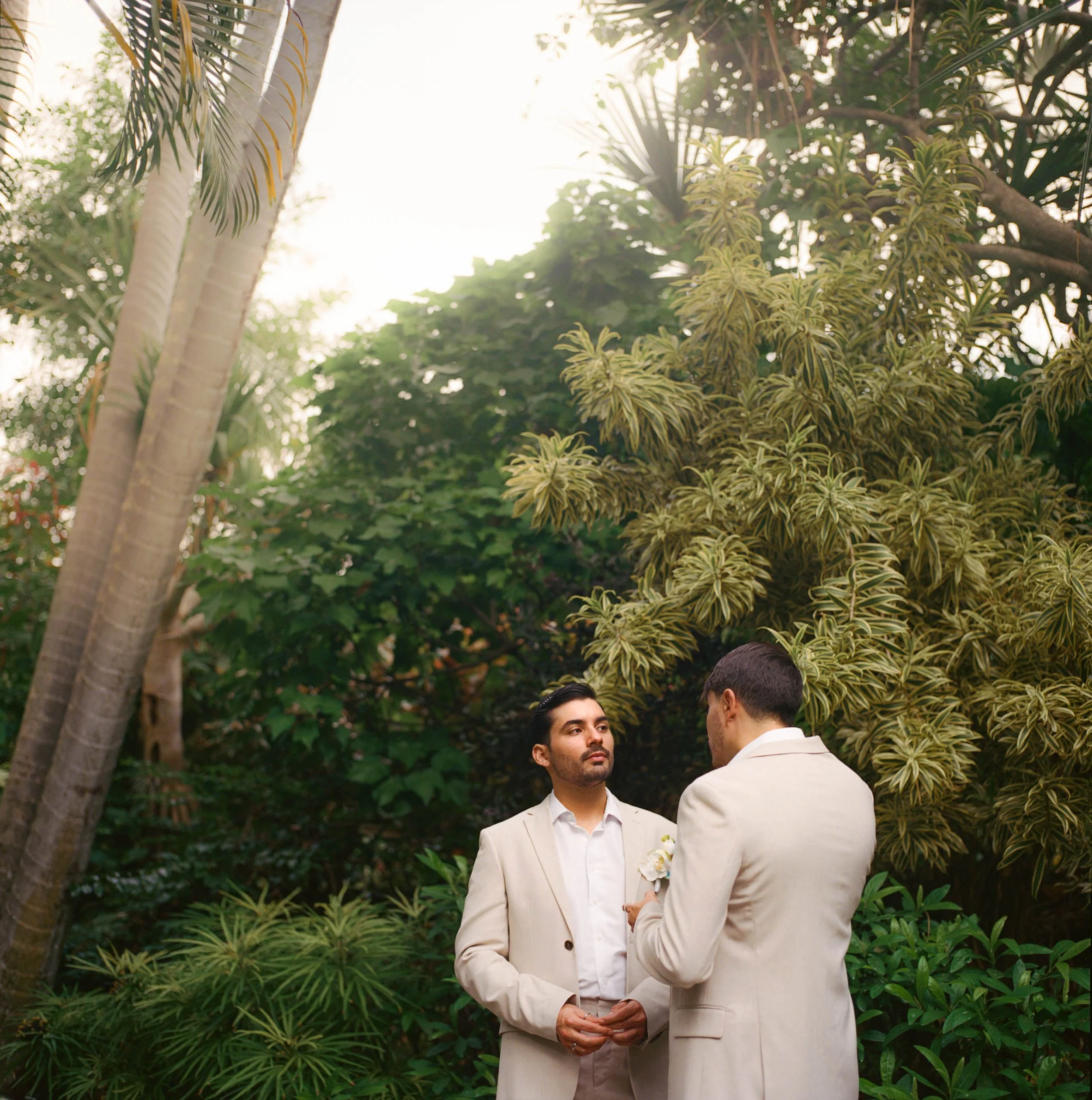 Two men in light-colored suits standing face-to-face outdoors among lush green trees, one holding a small object, with sunlight filtering through the leaves.