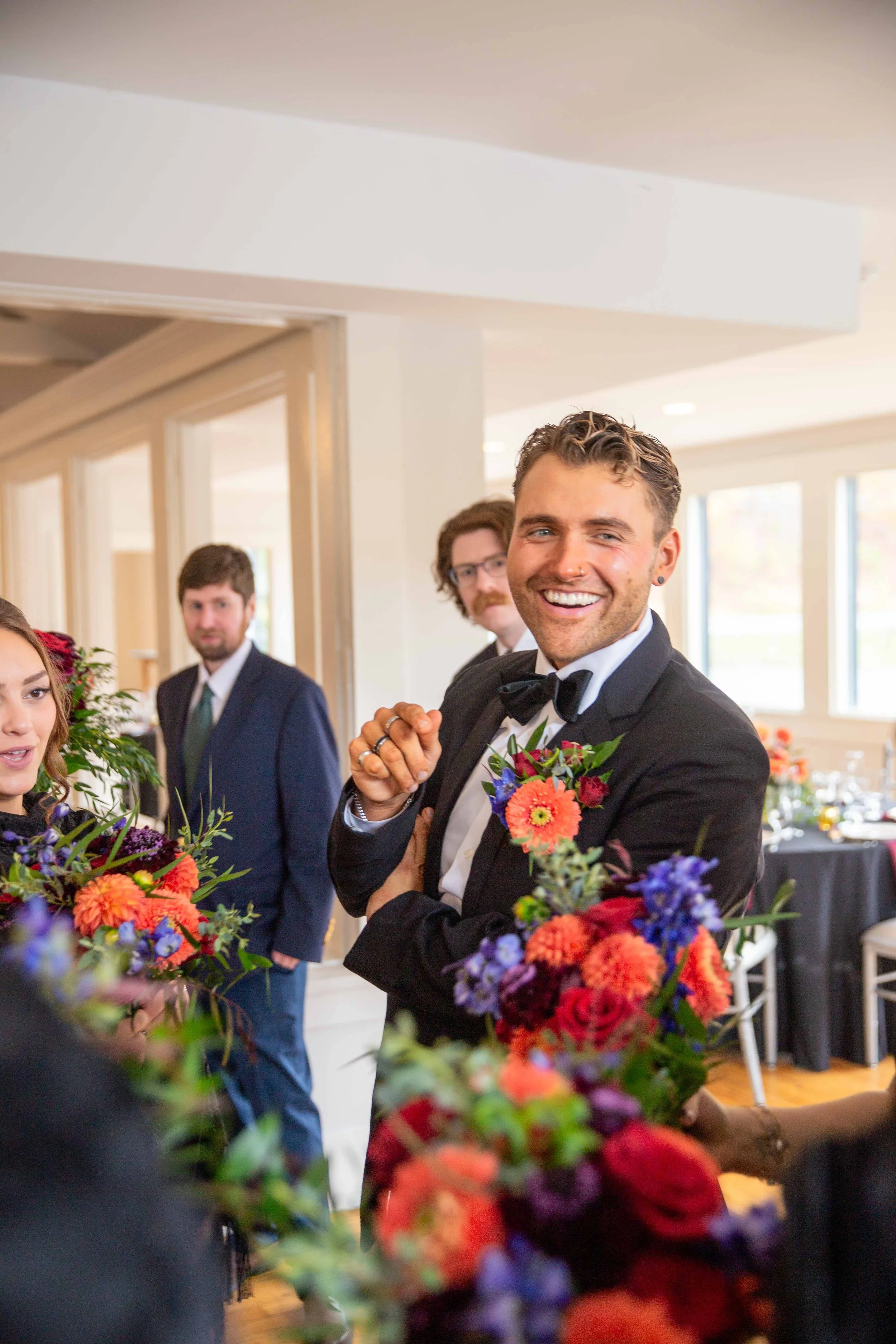 A smiling man in a tuxedo with a floral boutonniere at a wedding reception, surrounded by other guests and floral arrangements.