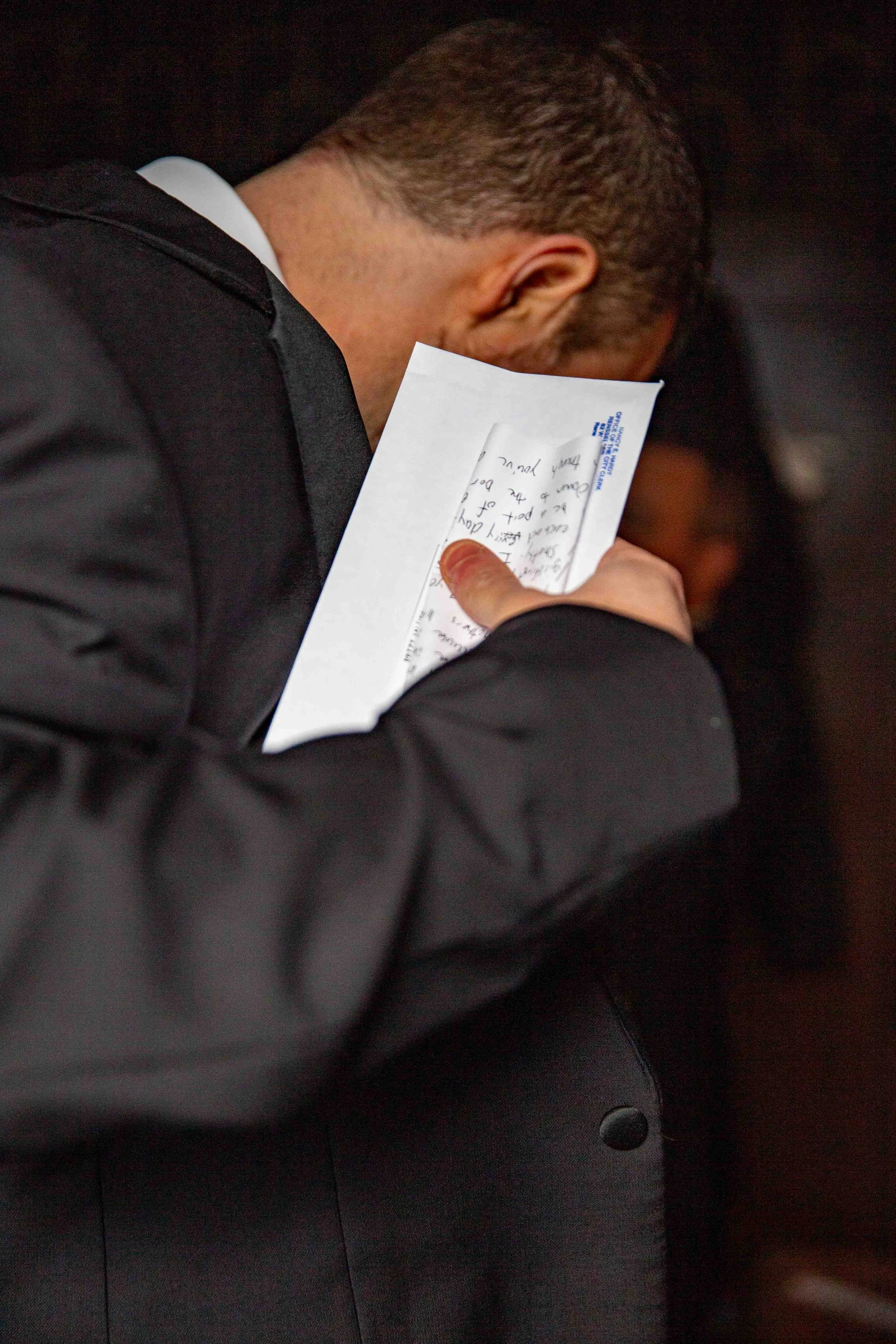 A man in a black suit leaning against a wall, holding a piece of paper or note close to his face and reading it.