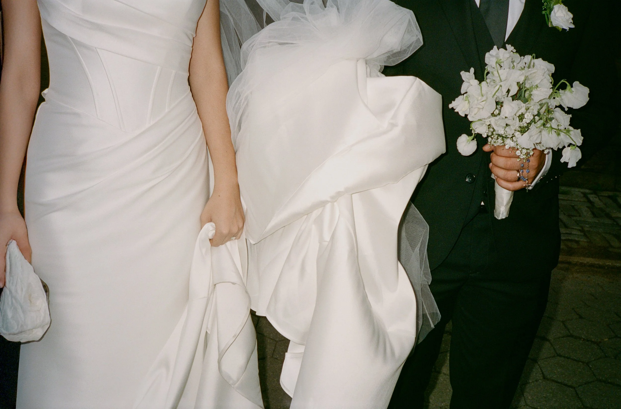 Close-up of a bride and groom at their wedding; the bride is holding her wedding dress, and the groom is holding a bouquet of white flowers.