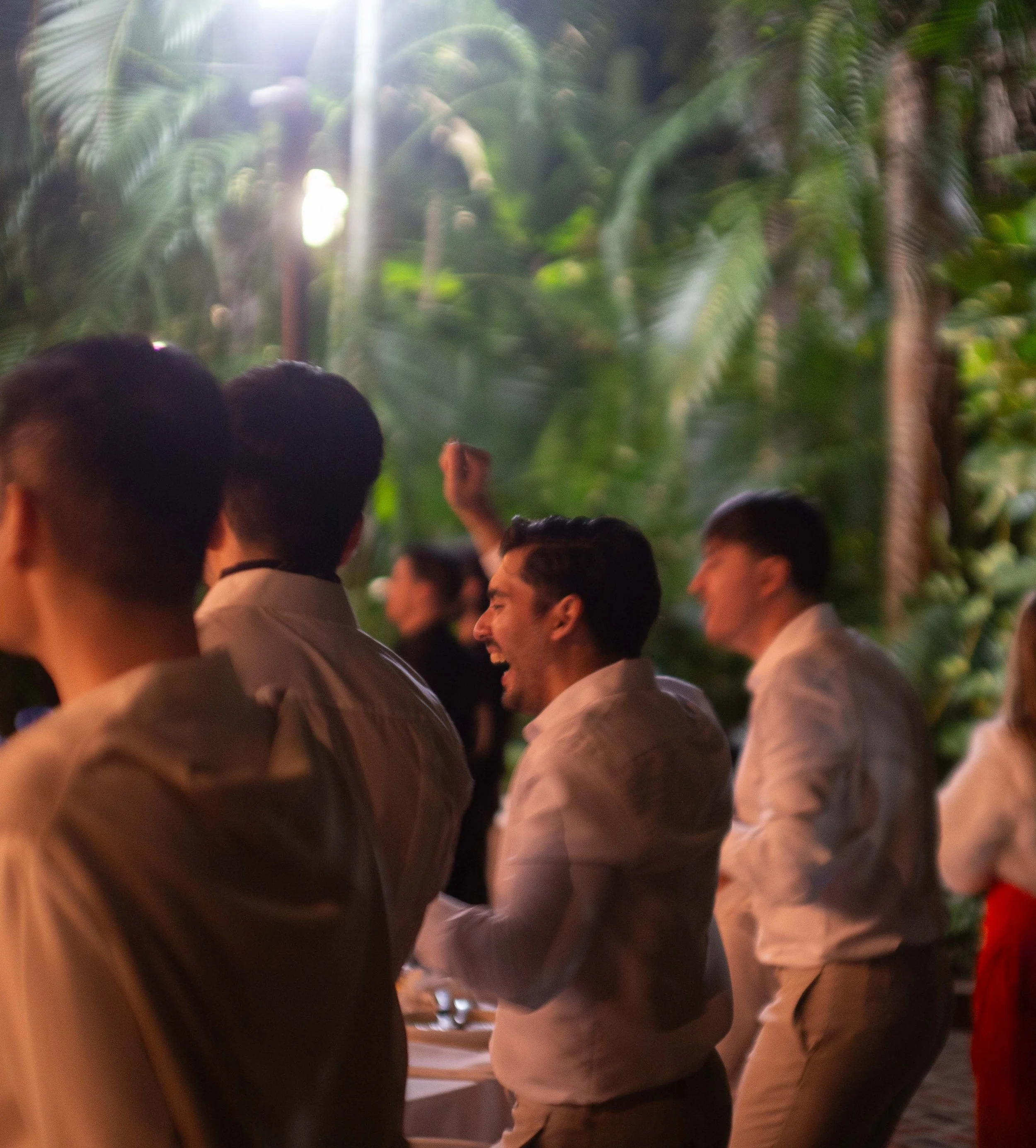 People enjoying an outdoor event at night, with lush green tropical plants in the background, some wearing light-colored shirts, one person laughing.
