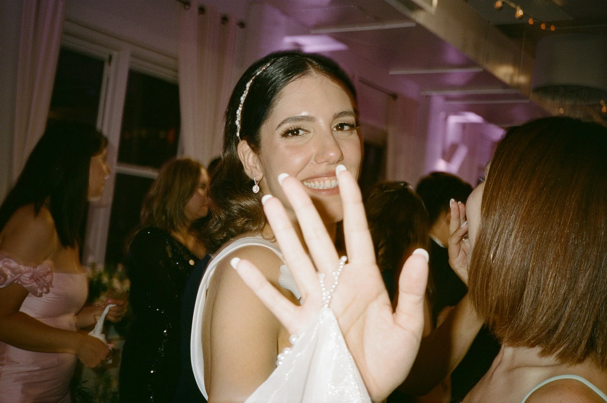 A woman with dark hair smiling and showing her hand with a white lace glove at a party or celebration, surrounded by other women in elegant dresses.