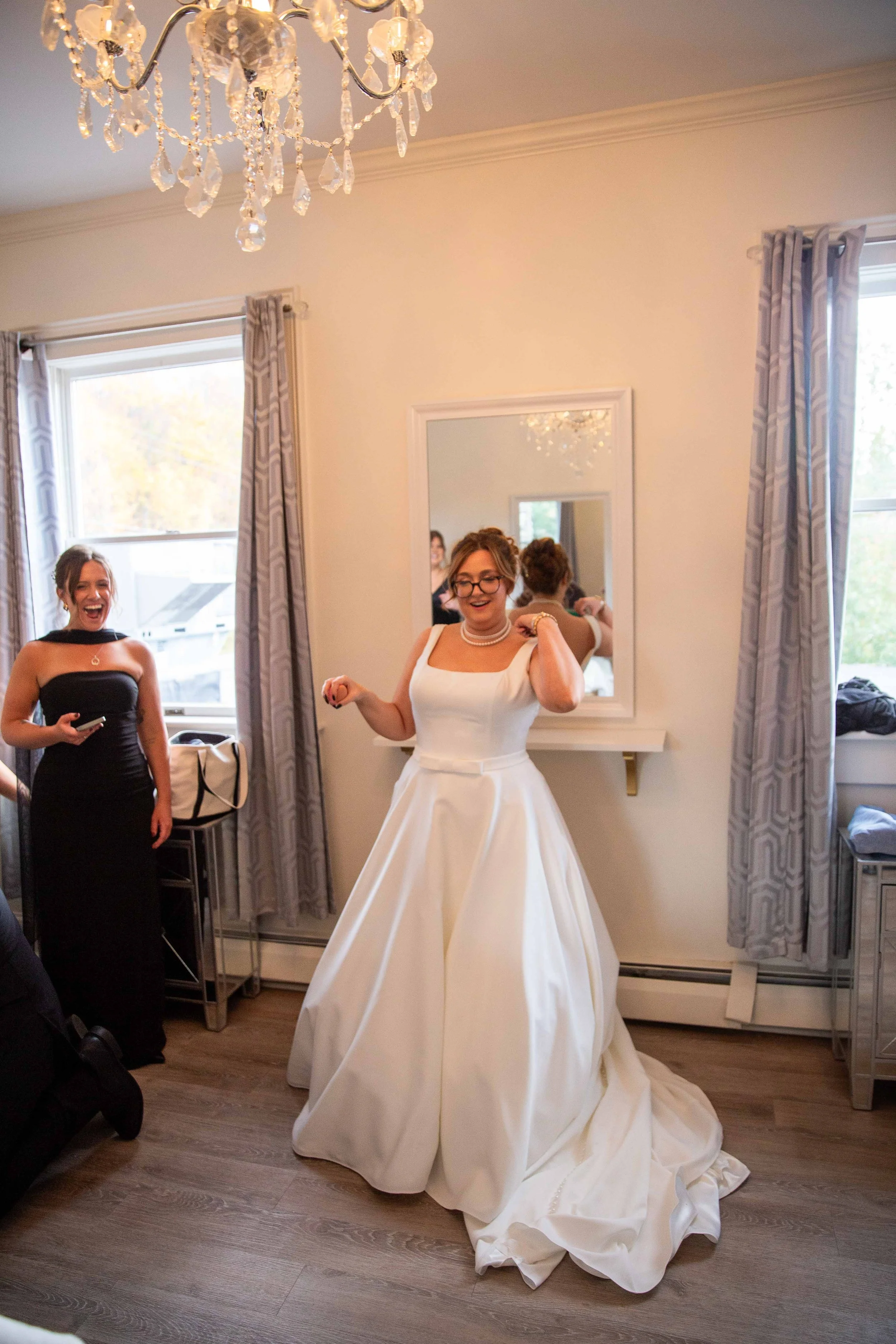 A bride in a white wedding gown smiling and adjusting her necklace inside a room with a chandelier and large windows, with a woman in a black dress laughing nearby.
