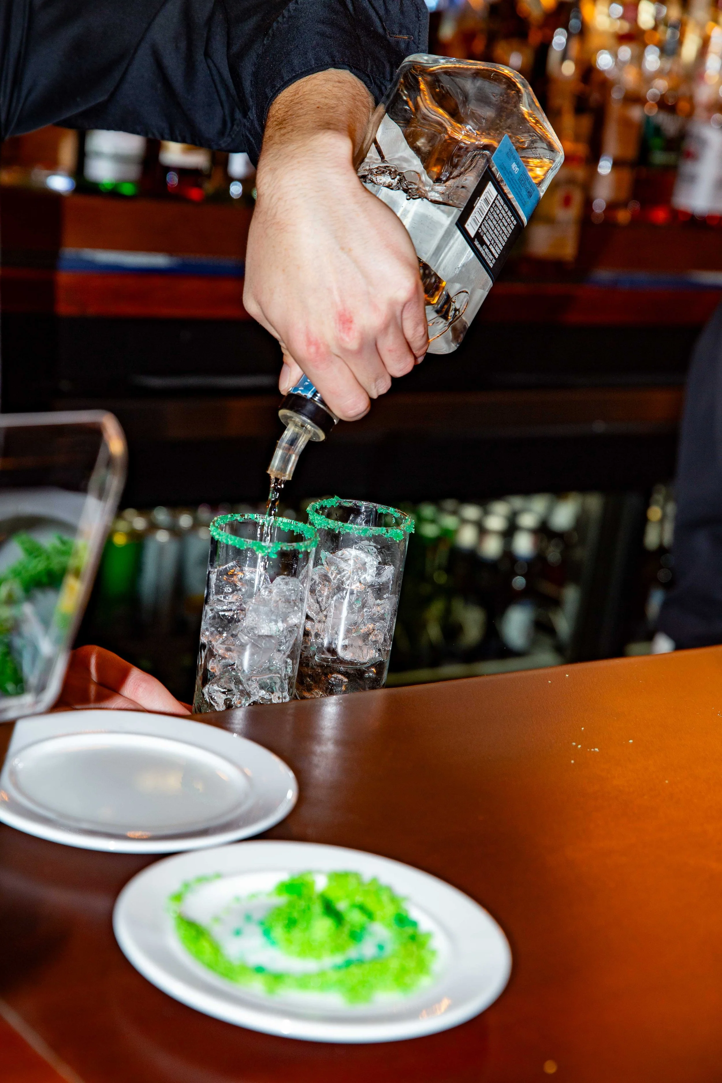 A person pouring whiskey into two glasses with ice, with green sugar-rimmed glasses on a bar counter, and a plate of green dessert in the foreground.