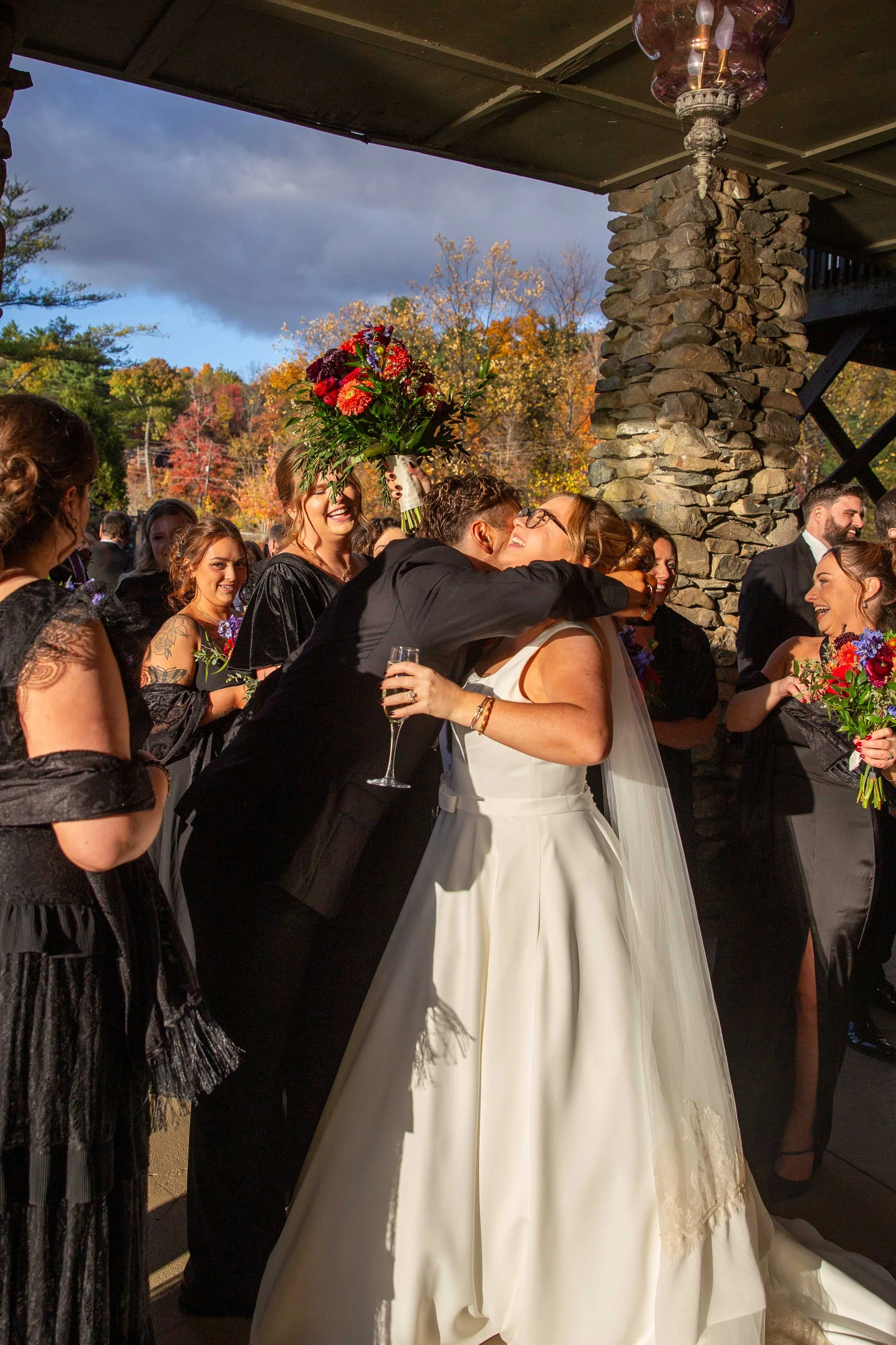A bride in a white wedding gown is hugging a man in a black suit, while the bridesmaids and guests in black dresses hold bouquets. One bridesmaid is holding a large bouquet of flowers above her head. The scene is outdoors with fall foliage in the bac