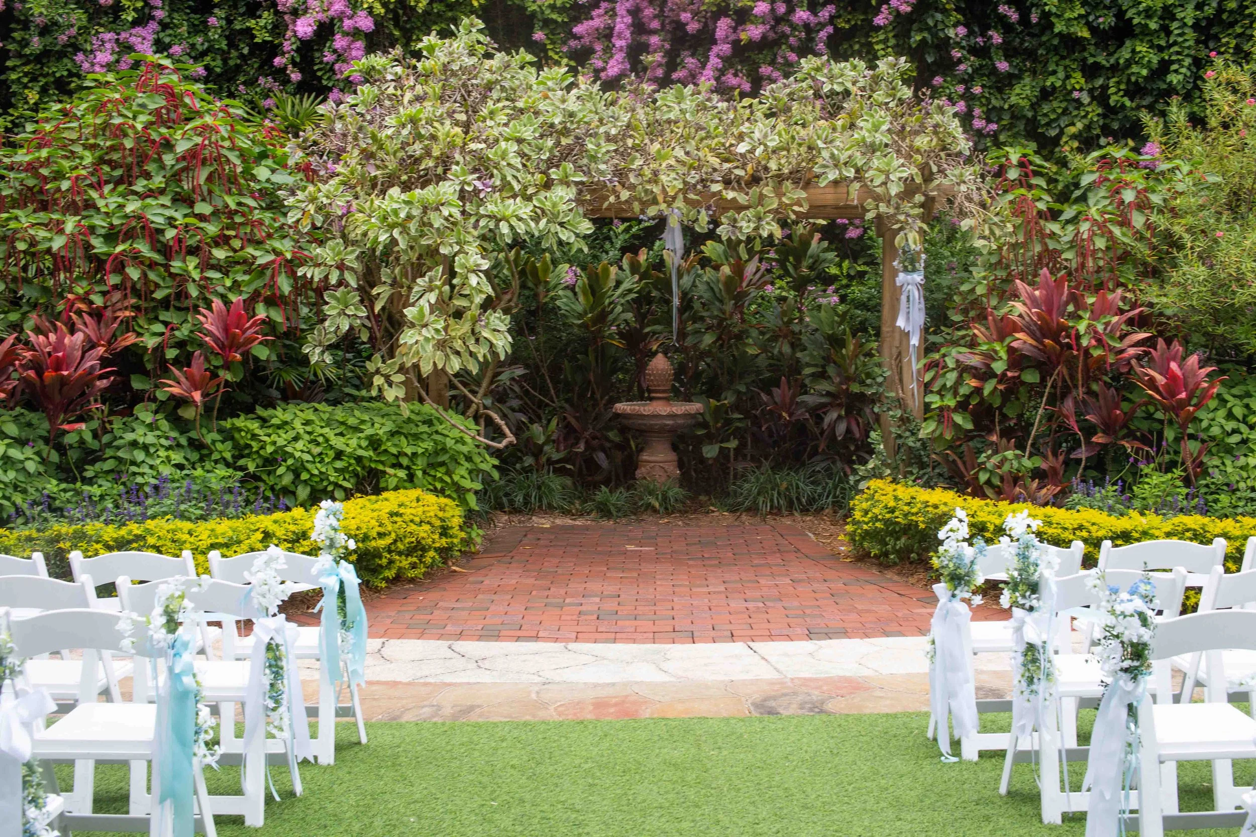 Outdoor wedding ceremony setup with white chairs decorated with flowers and ribbons, a brick pathway leading to a garden altar with a fountain surrounded by lush green plants and colorful flowers.