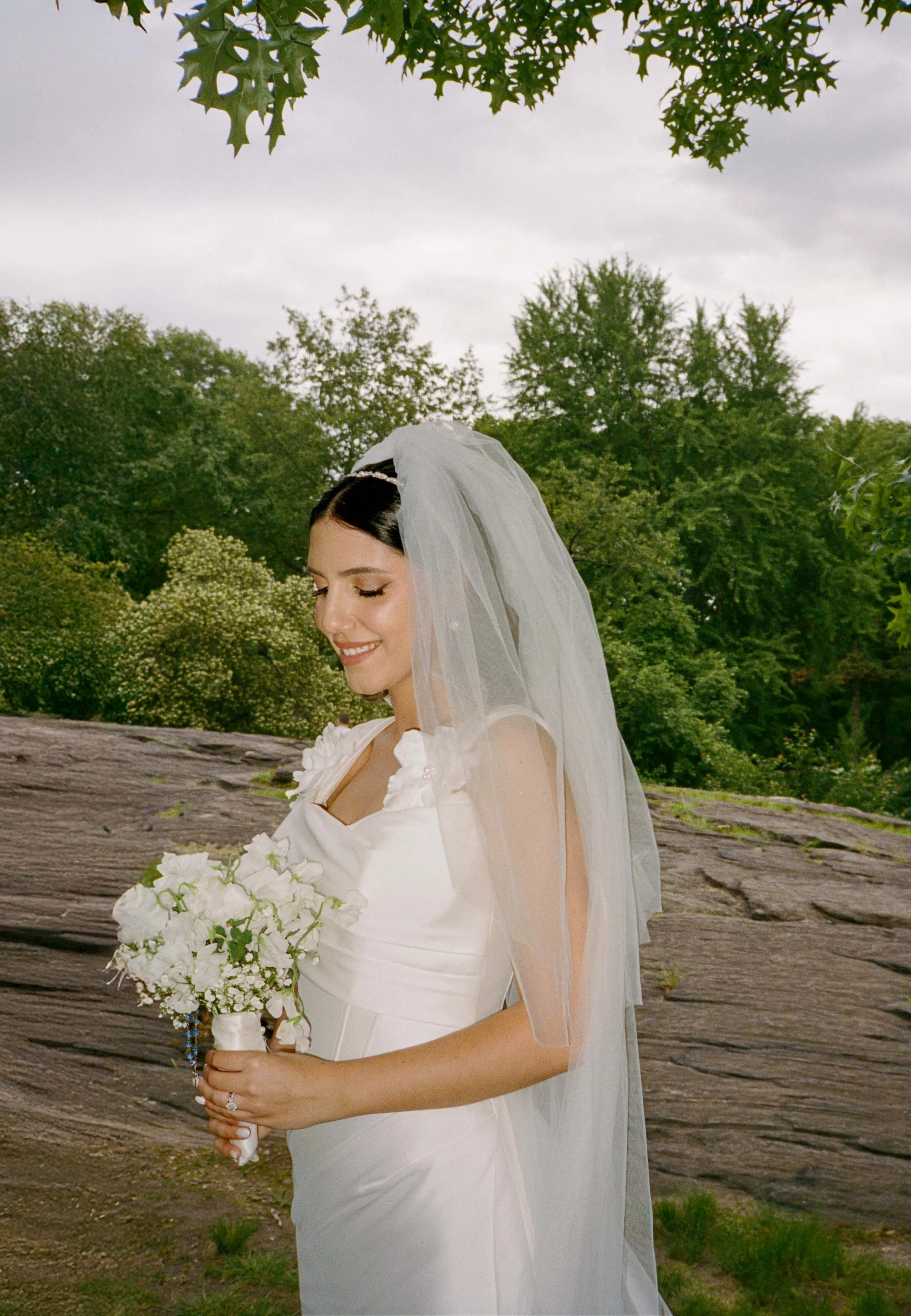 A bride in a white wedding dress with floral embellishments, holding a bouquet of white flowers, and smiling with her eyes closed outdoors with green trees and a stone surface background.