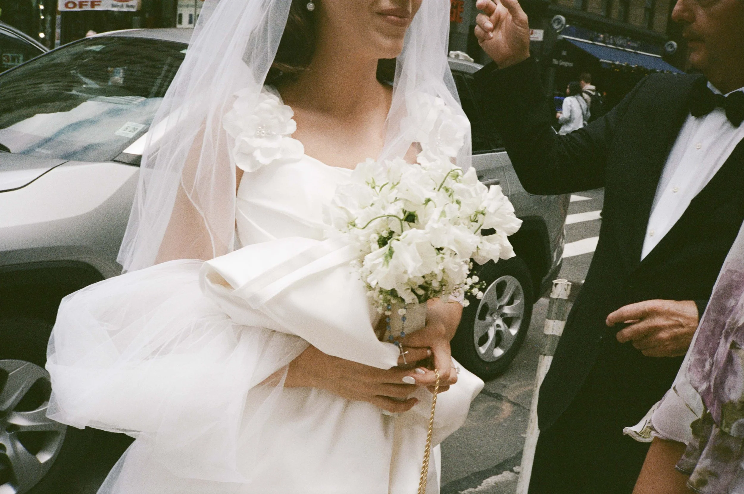 A bride in a white wedding dress holding a bouquet of white flowers, standing on a city street with a man in a black tuxedo.