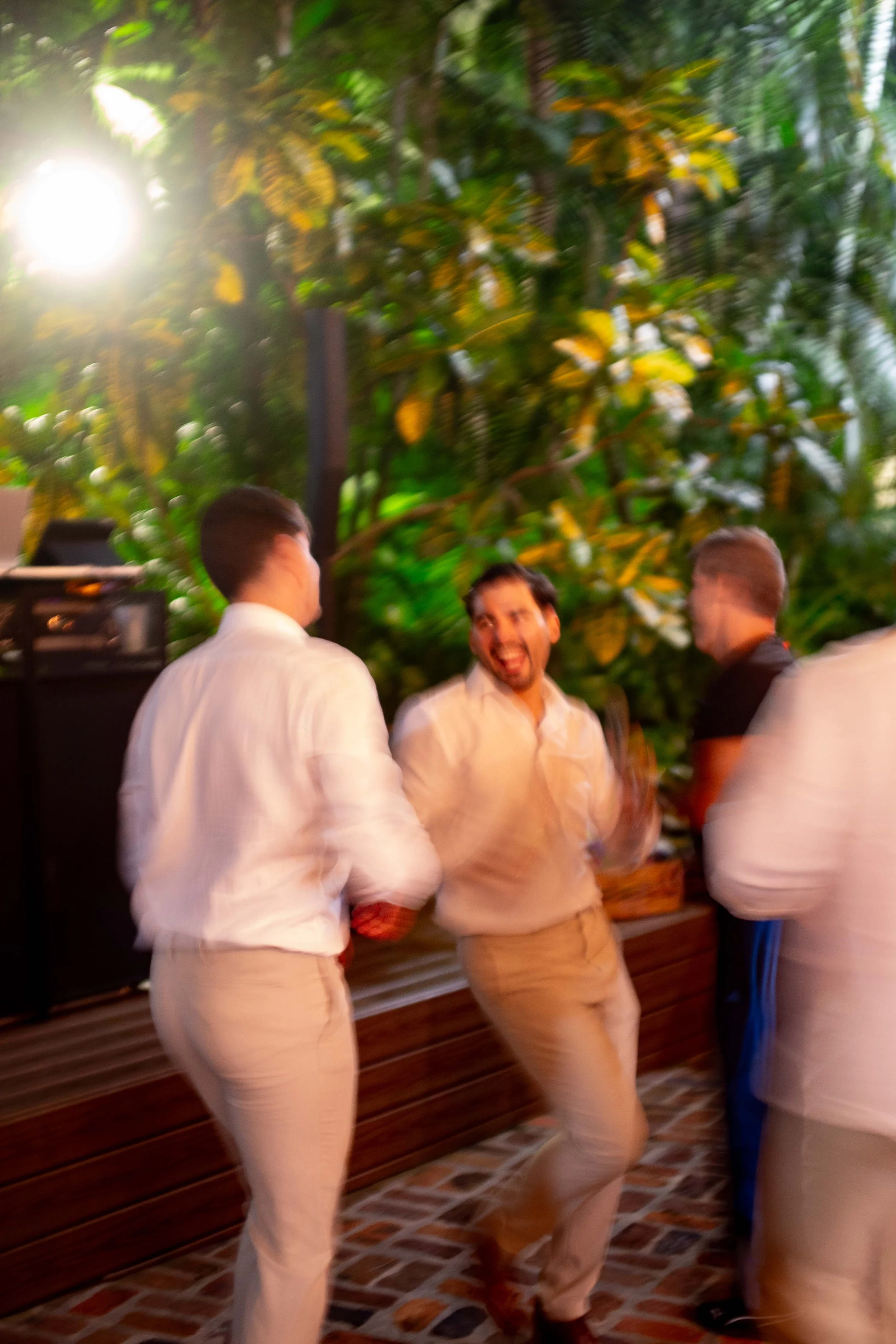 Three men are dancing and enjoying themselves at an outdoor event, with lush green plants and warm lighting in the background.