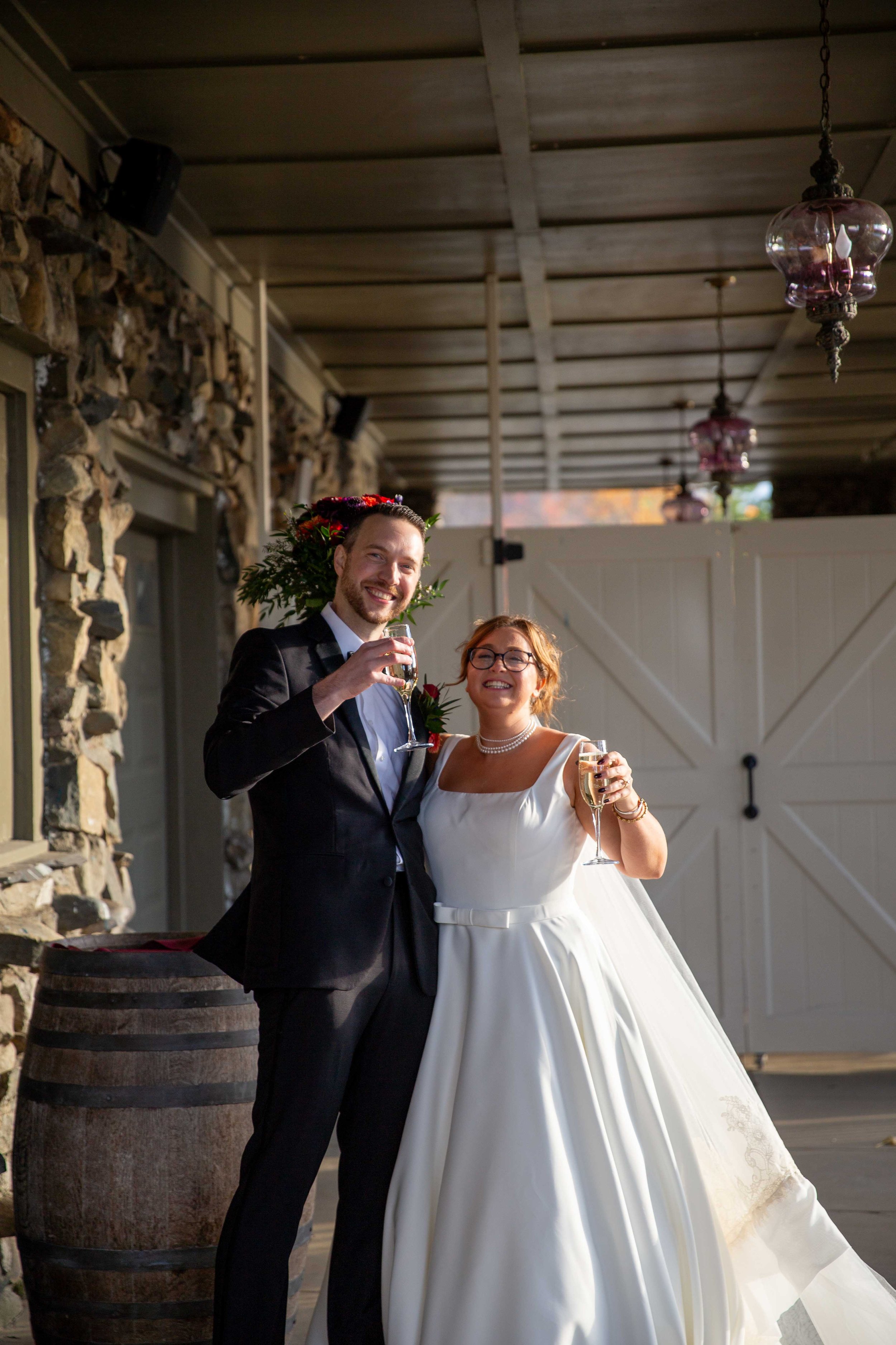 A newlywed couple celebrating with glasses of champagne on a rustic outdoor patio with stone walls, wooden ceiling beams, and hanging pink glass lanterns.