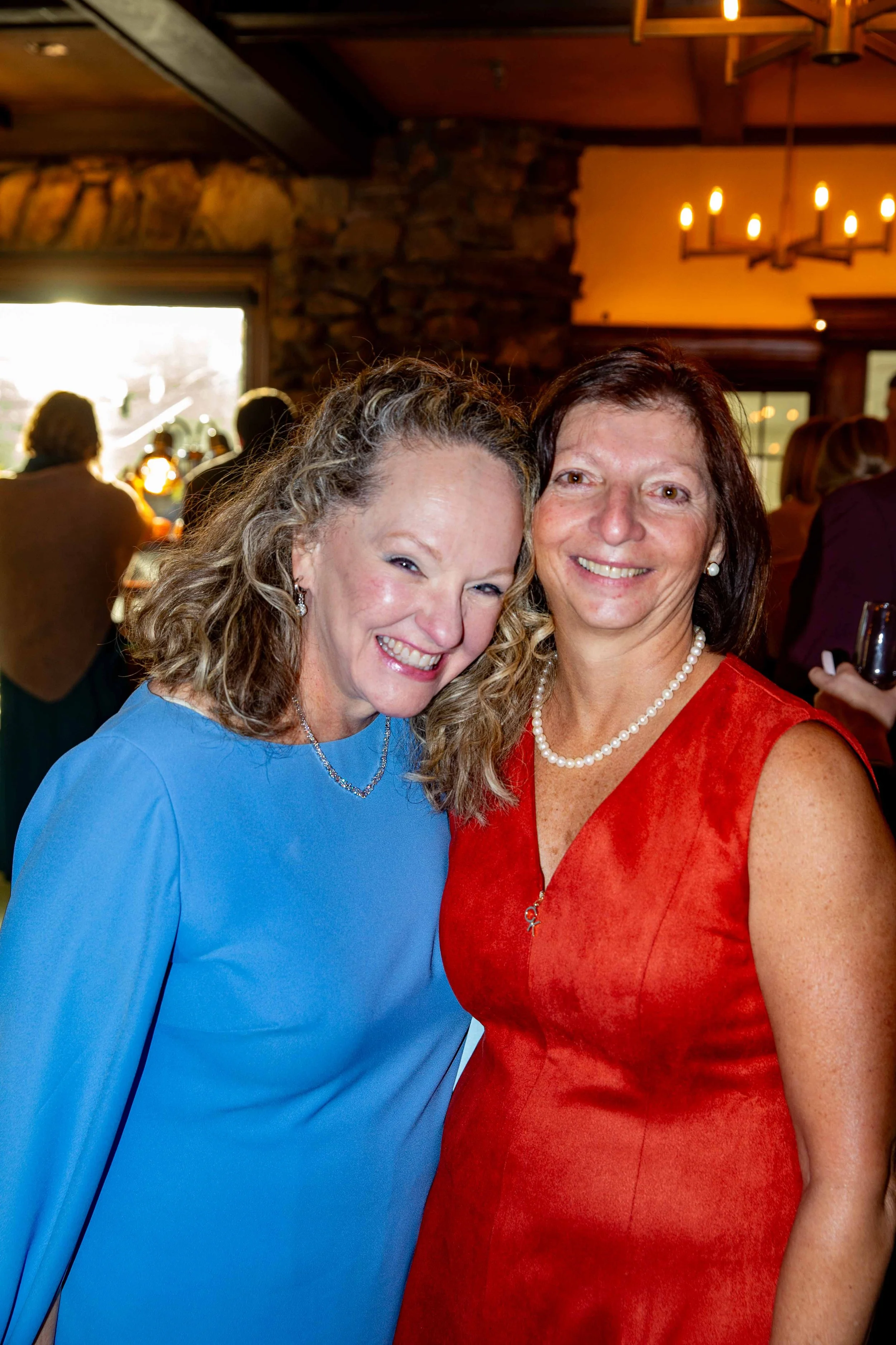Two women smiling, one in a blue dress and the other in a red dress, at a social gathering indoors.
