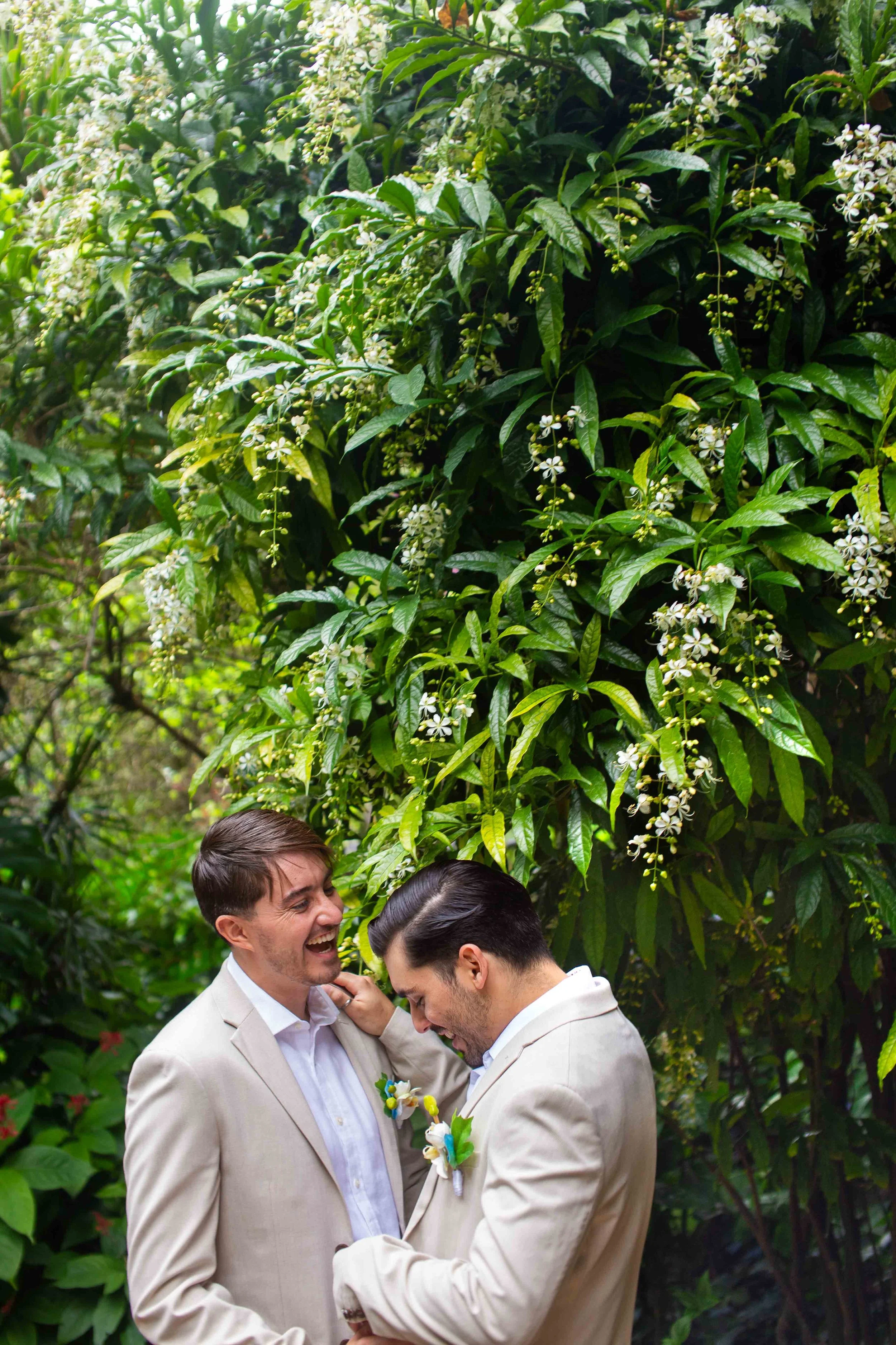Two men in beige suits sharing a joyful moment in front of lush green plants with white flowers.