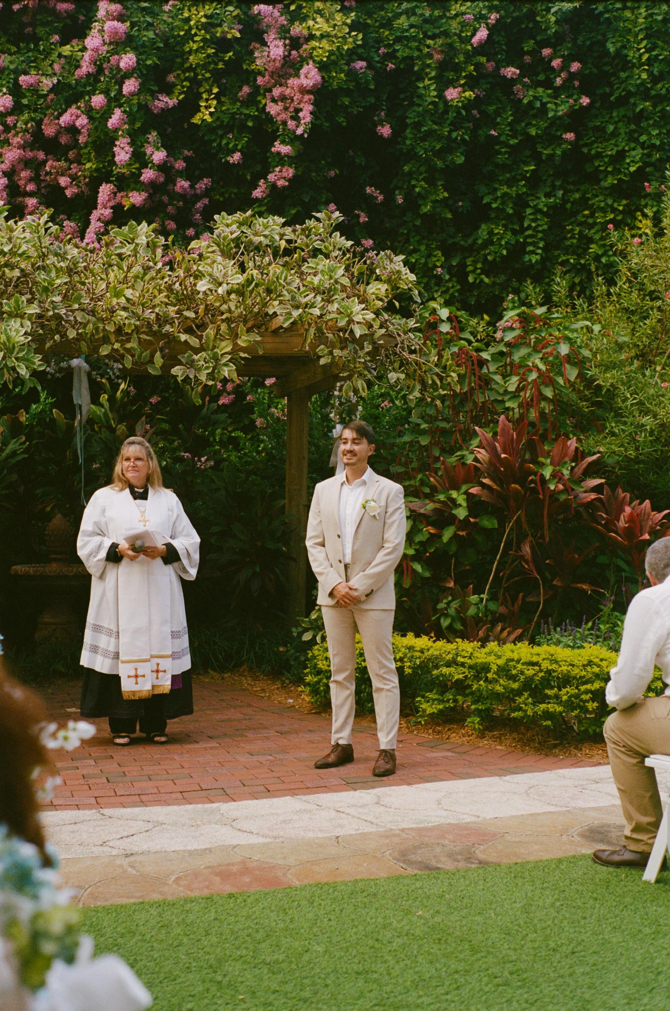 A wedding ceremony taking place outdoors in a lush garden with vibrant greenery and pink flowers; a woman in religious attire officiates, a man in a light-colored suit stands with a smile, and another man kneels in front of them.