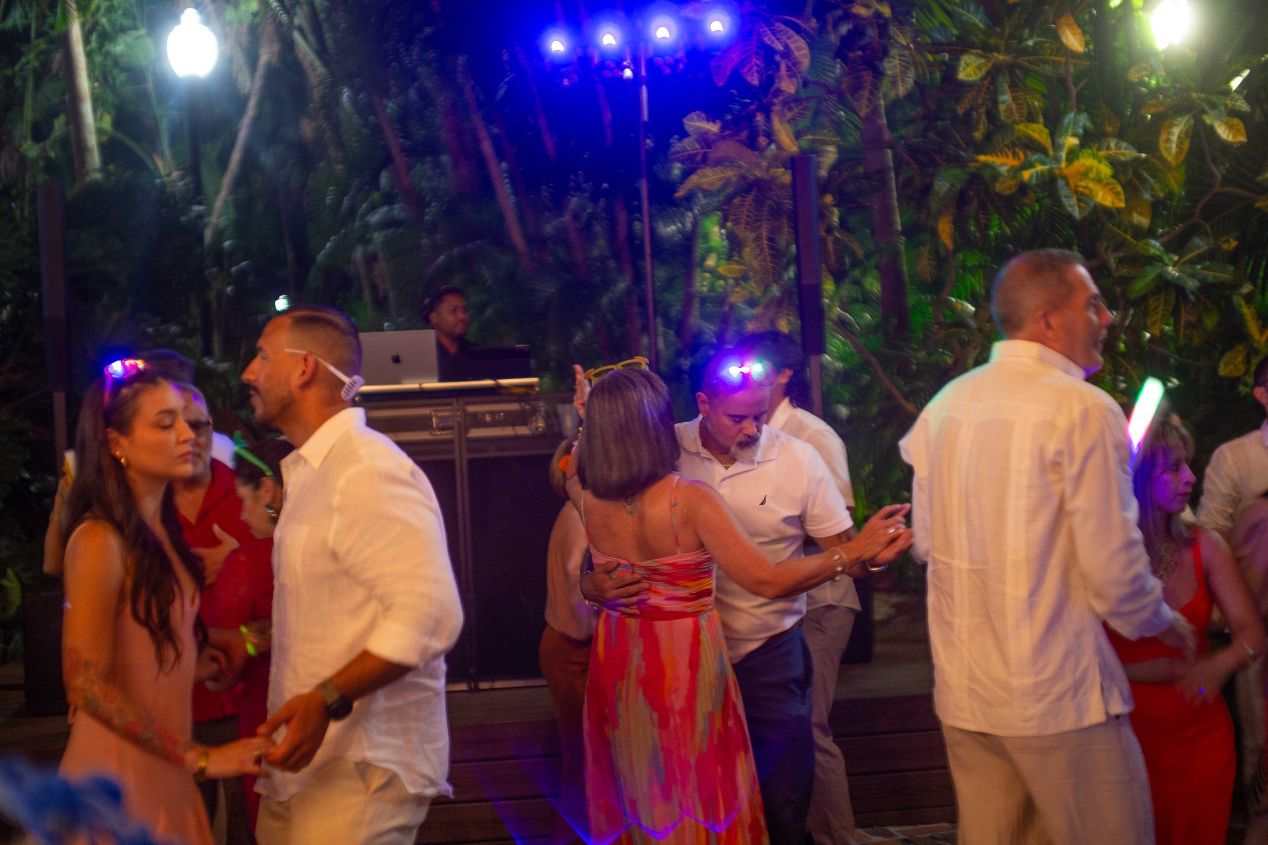 People dancing at a party, with a DJ in the background and tropical plants surrounding the scene.