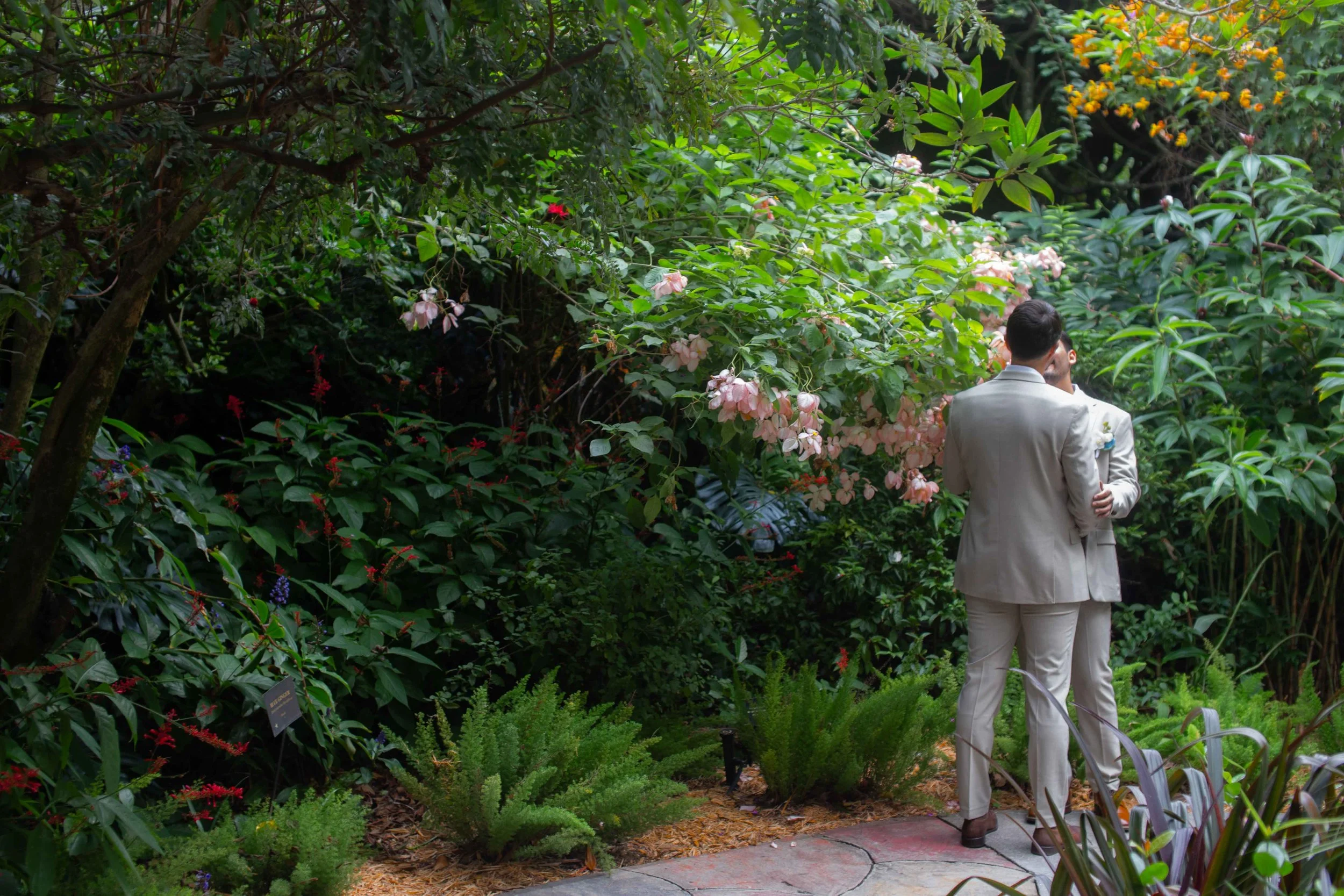 Two men dressed in light-colored suits embracing in a garden surrounded by lush green foliage and pink flowering bushes.