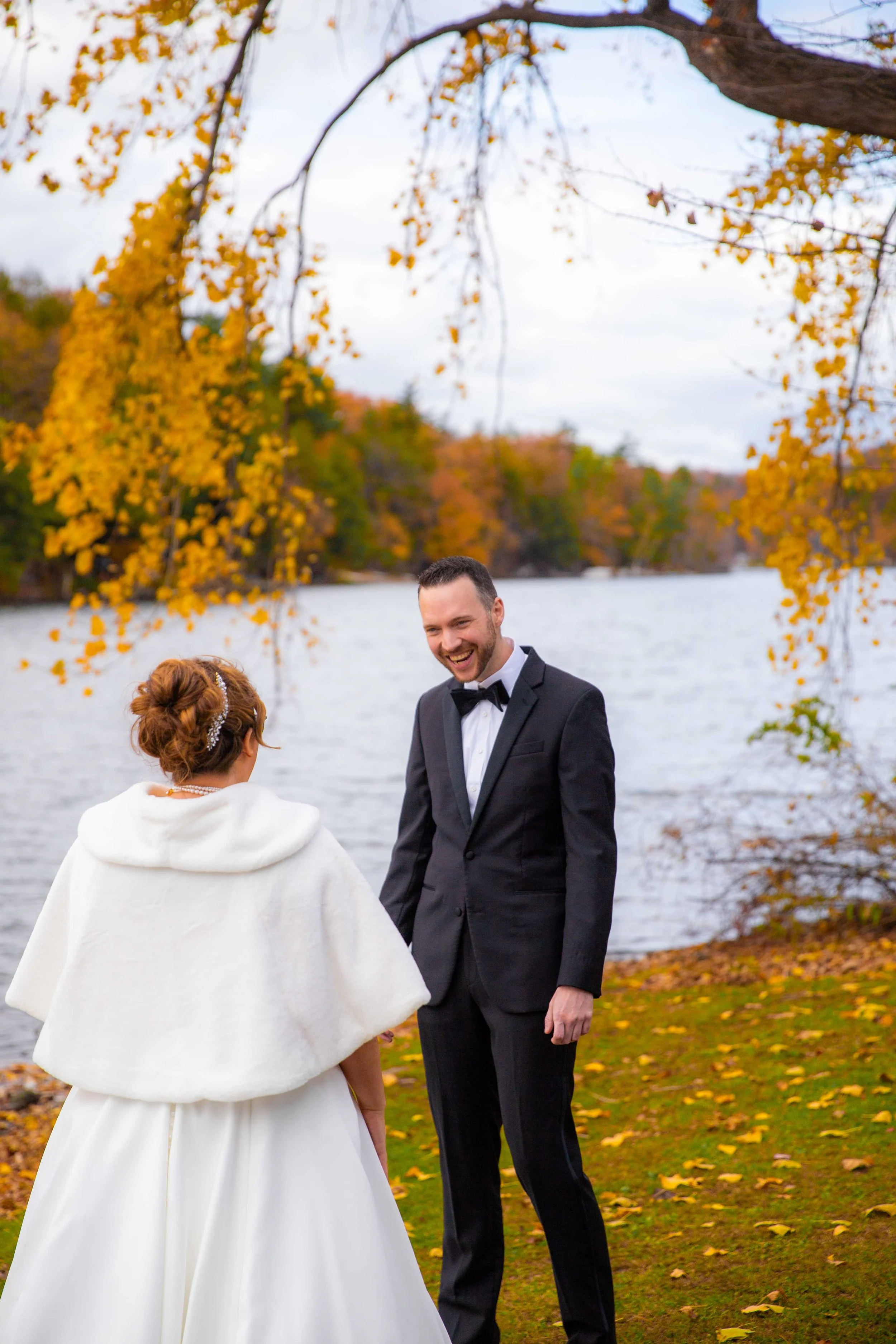 A bride and groom smiling at each other outdoors by a lake during autumn, with yellow leaves on trees and fallen on the ground.
