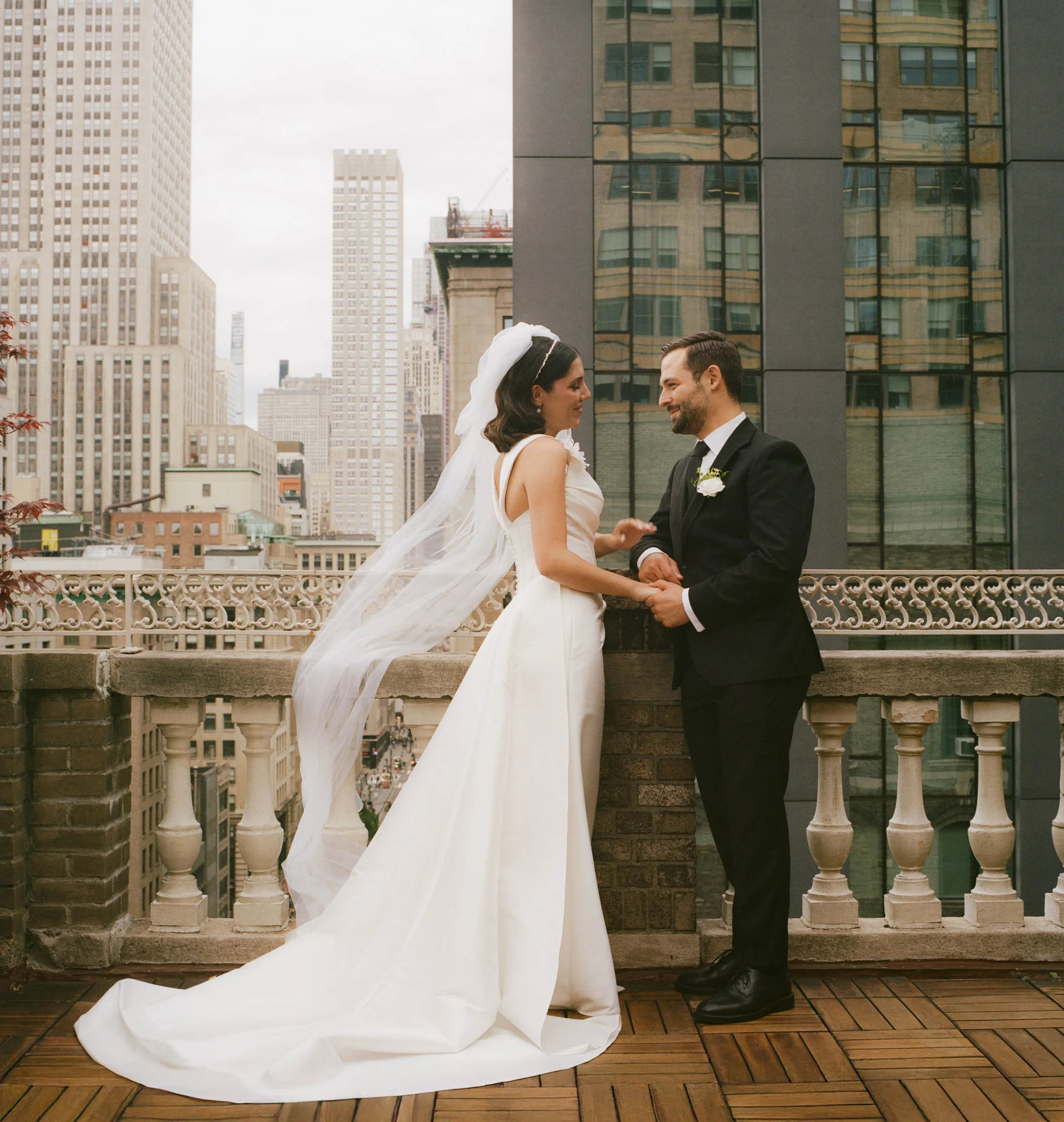 A bride and groom hold hands and stare into each other's eyes on a city rooftop, with tall buildings in the background.