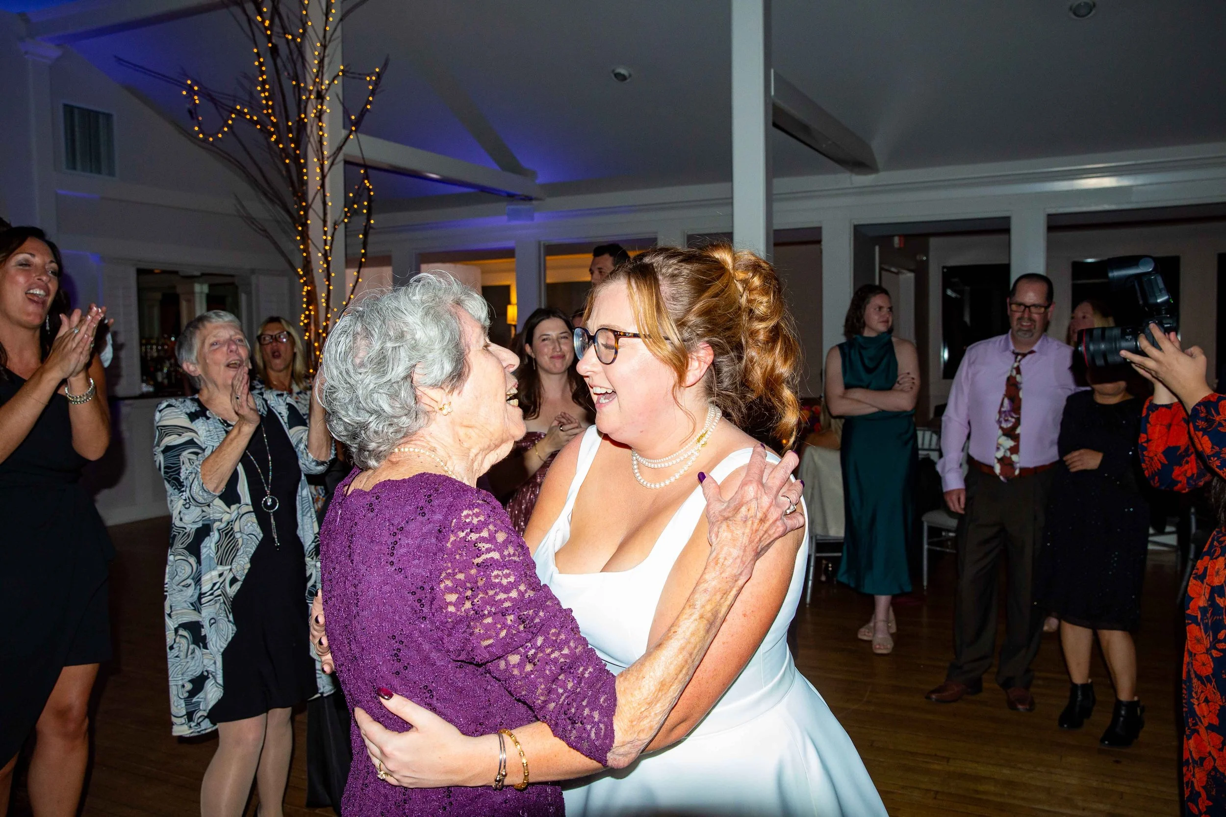 A bride in a white dress with glasses and pearl jewelry dances with an elderly woman in a purple dress at a celebration party. Guests in the background are clapping and enjoying the moment.
