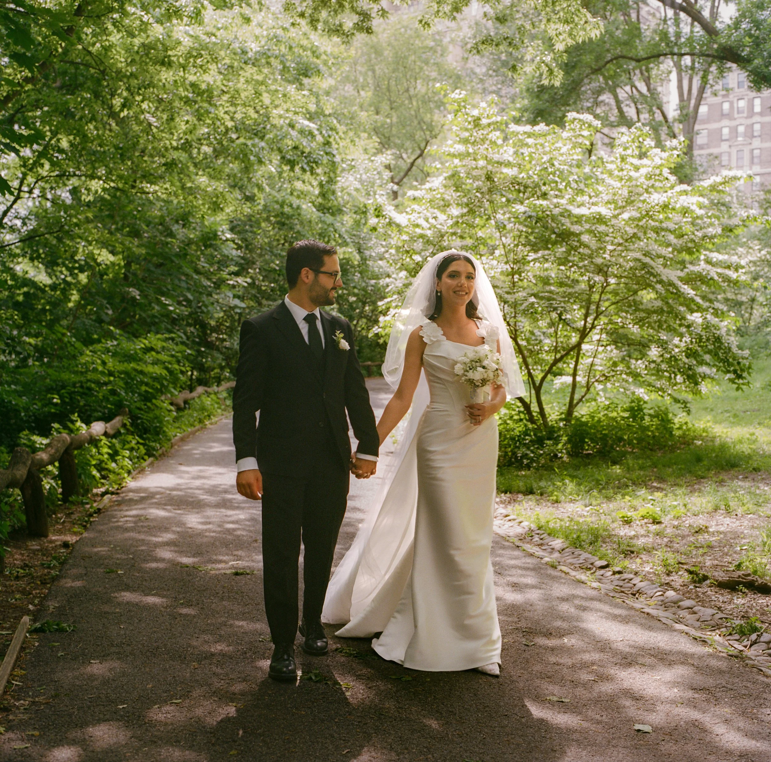 A bride and groom walking hand in hand on a park pathway surrounded by lush green trees, with the bride holding a bouquet of white flowers and wearing a white wedding gown with a veil, while the groom is in a black suit, white shirt, and black tie.