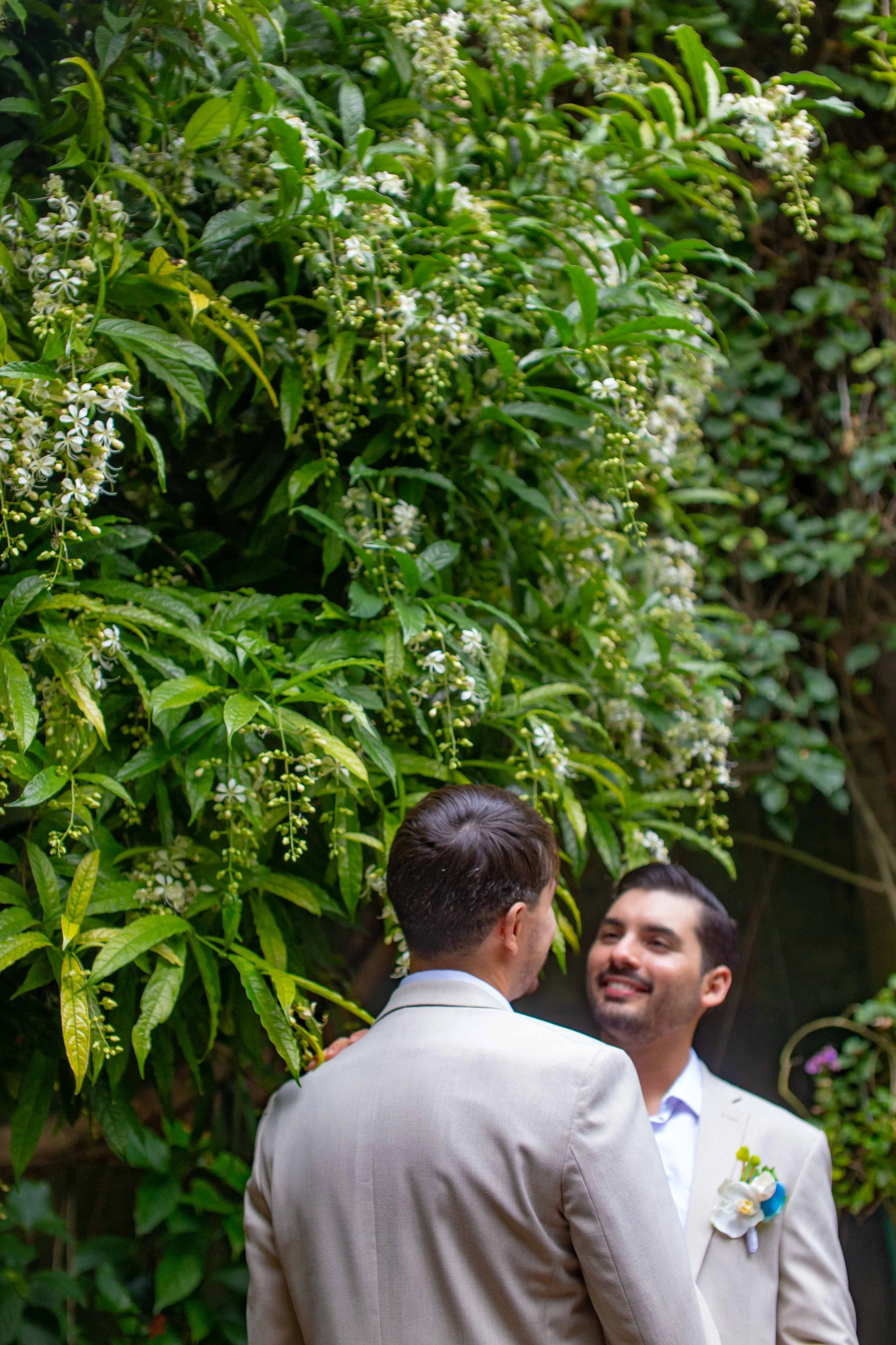 Two men in suits facing each other and smiling outdoors in front of lush green foliage with white flowers.