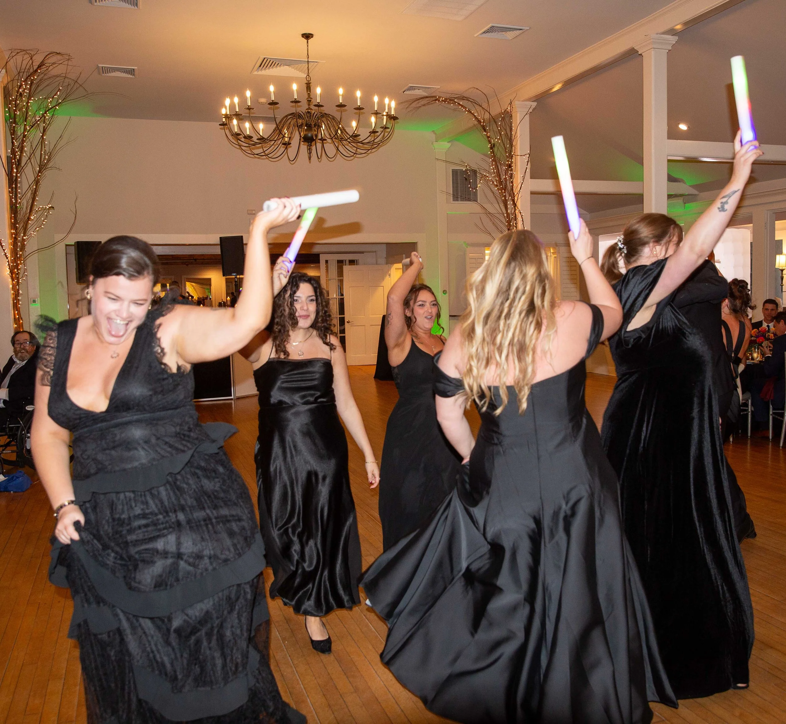 Women dancing at a party in elegant black dresses, holding glowing sticks, with some smiling and enjoying the music.