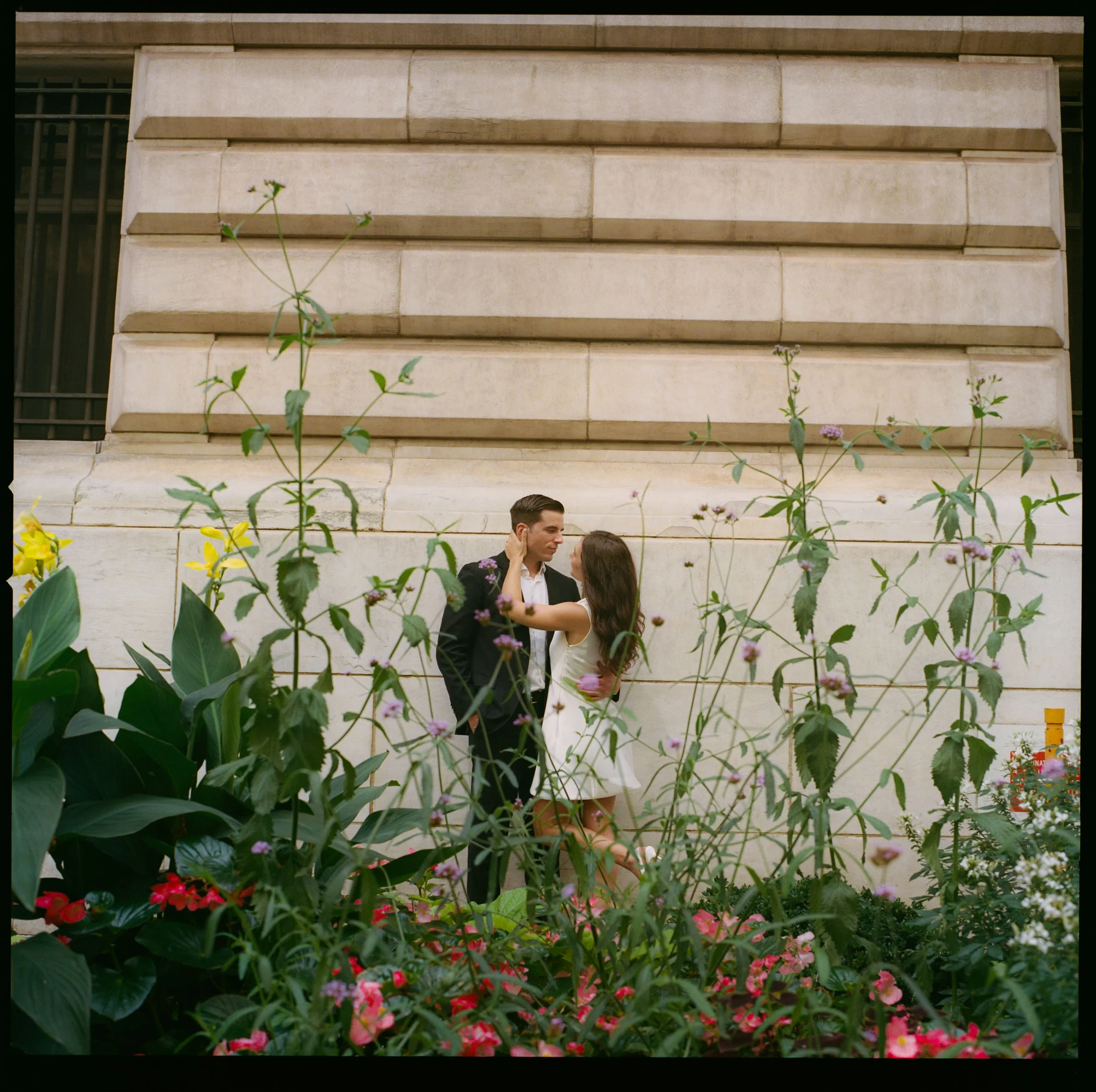 A couple standing closely face to face, surrounded by flowers, with a stone wall and window behind them.