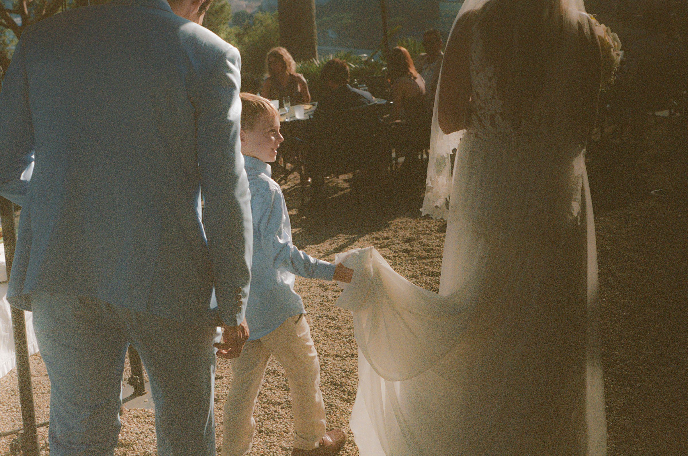 A young boy holding hands with a man in a light gray suit and a woman in a wedding dress during an outdoor wedding ceremony, with guests seated at tables in the background and trees in the distance.