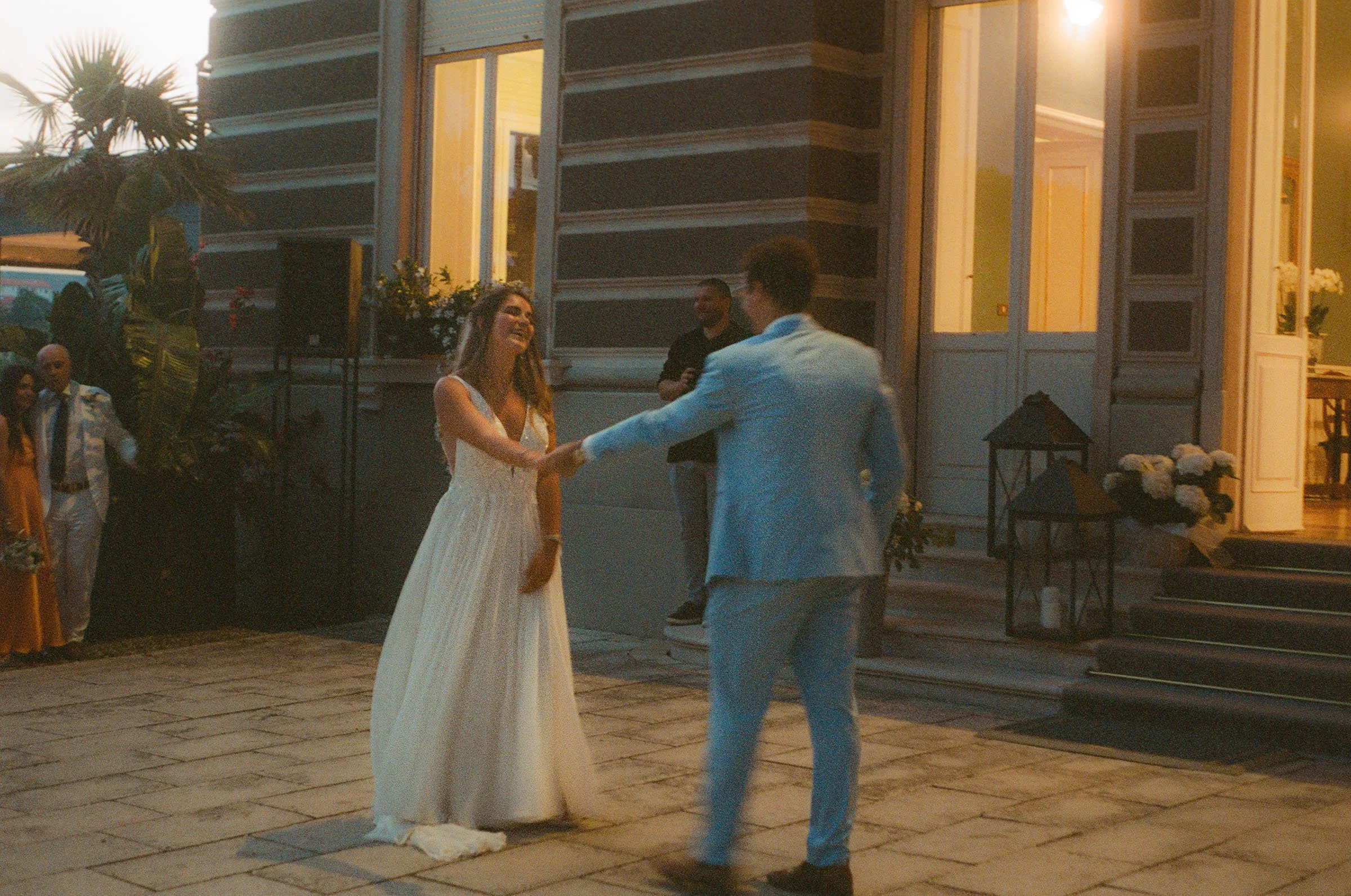 A couple dancing and holding hands outside a building during an evening event, surrounded by guests and warm lighting.