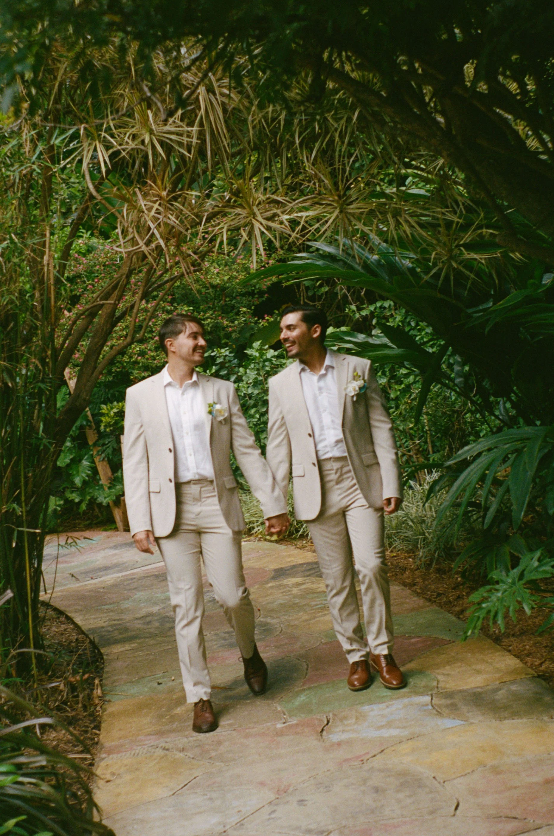 Two men in light-colored suits holding hands and smiling, walking on a stone path surrounded by lush green plants in a tropical garden.