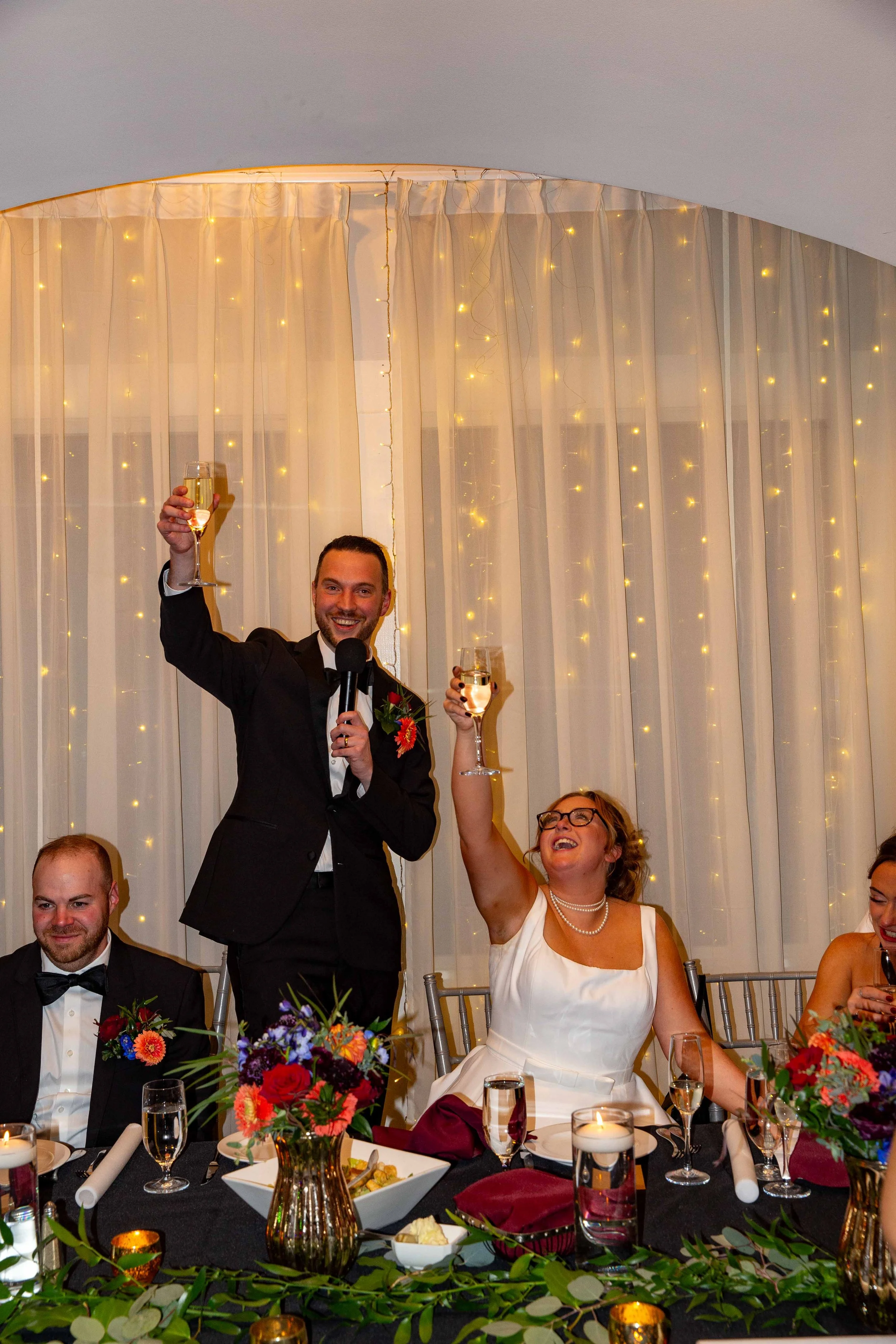 A group of people celebrating at a wedding reception, with the bride and groom raising glasses of champagne. The groom is standing, holding a microphone, and the bride is seated, smiling and wearing glasses. The background features white curtains wit