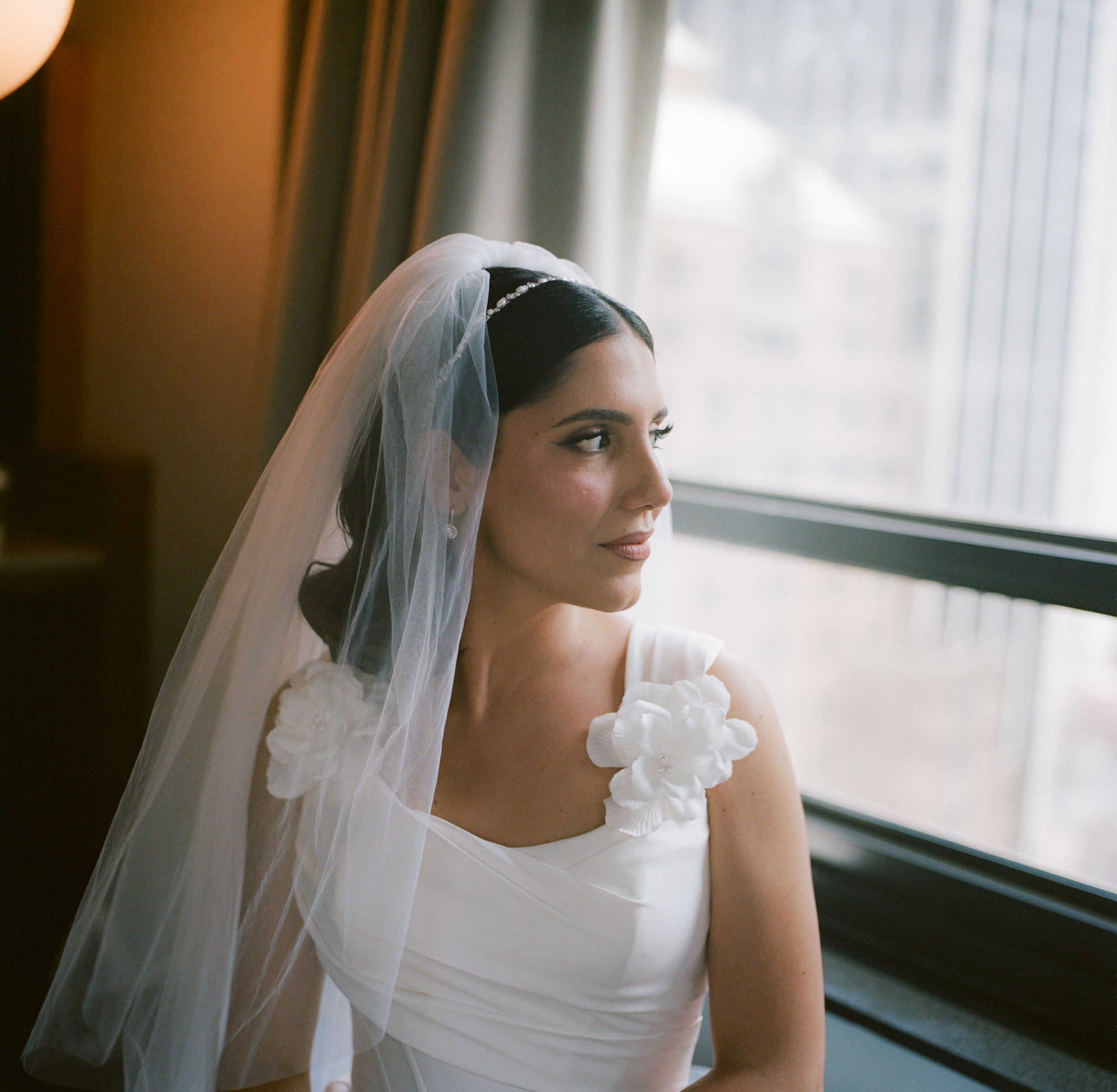 A bride standing by a window, looking outside, wearing a white wedding dress with 3D floral embellishments on the shoulder and a veil.