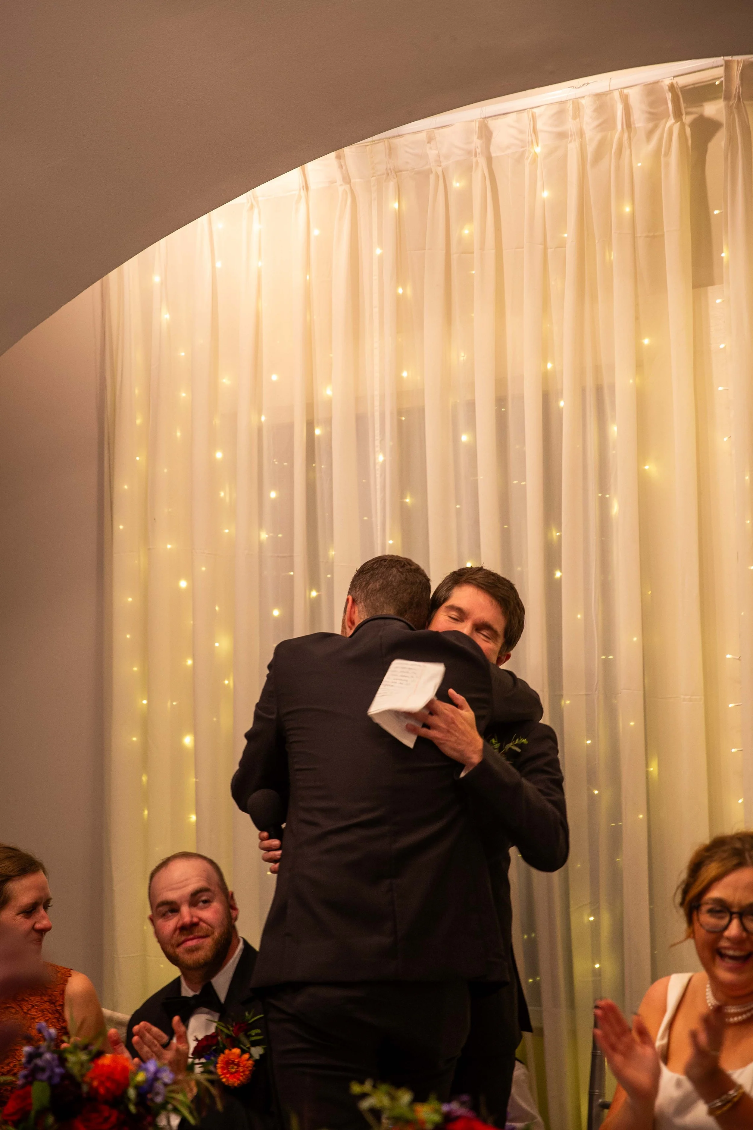 Two men hugging, one holding a microphone, during what appears to be an emotional celebration or speech, with other guests clapping and smiling at a decorated event.