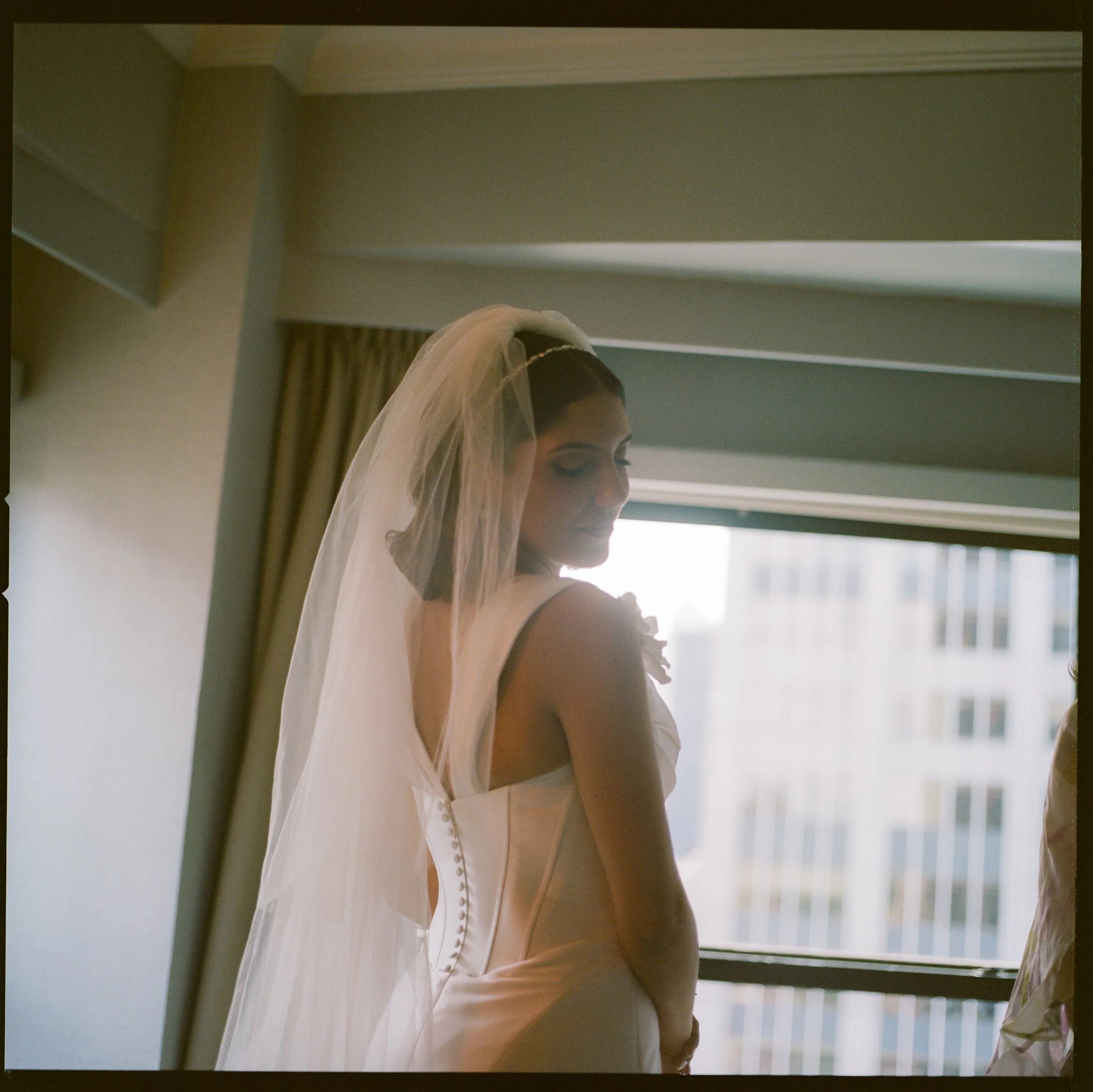 A woman in a wedding dress with a veil standing indoors near a window.