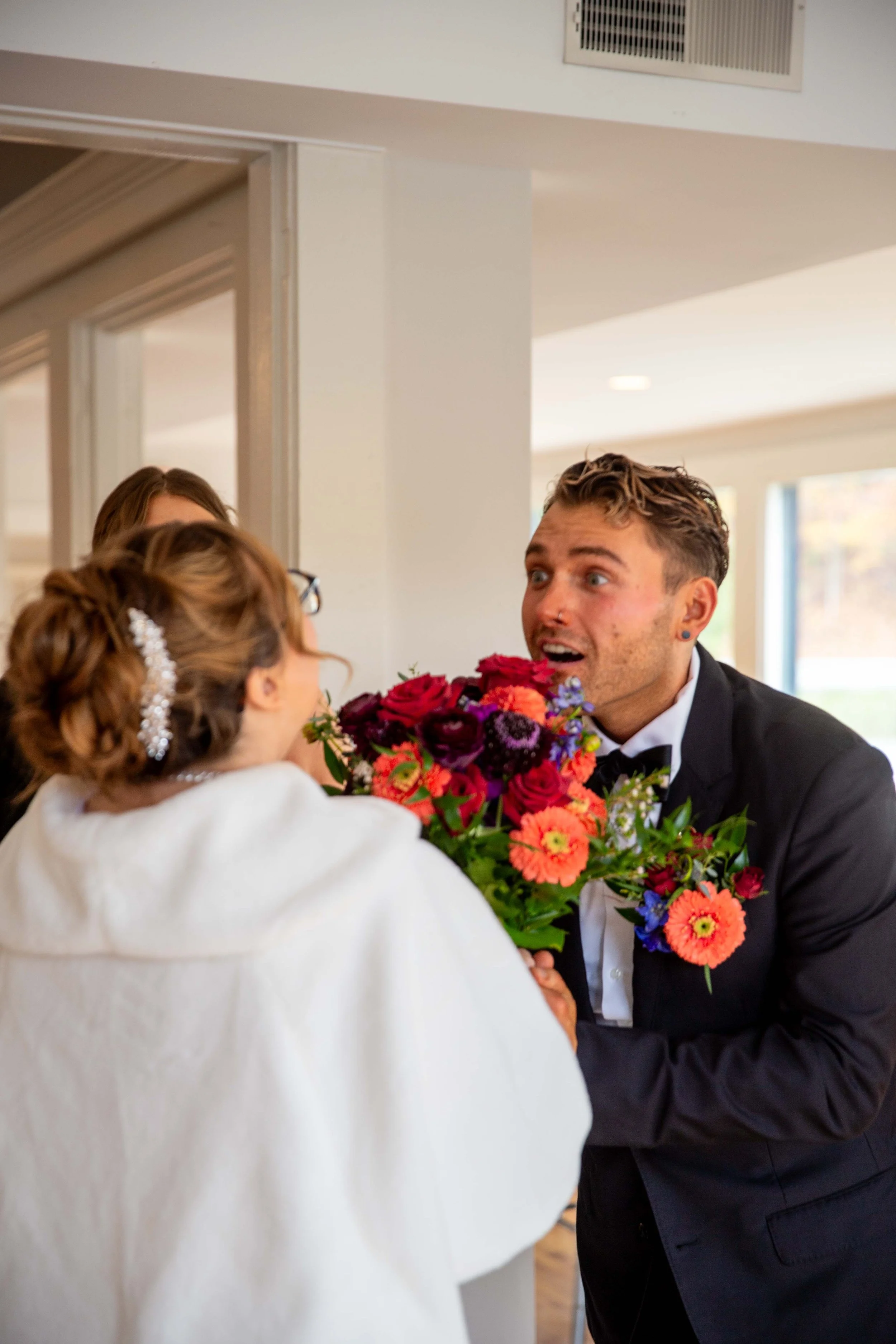 A man in a tuxedo offers a bouquet of colorful flowers to a woman dressed in white, with other women in the background, in an indoor setting.
