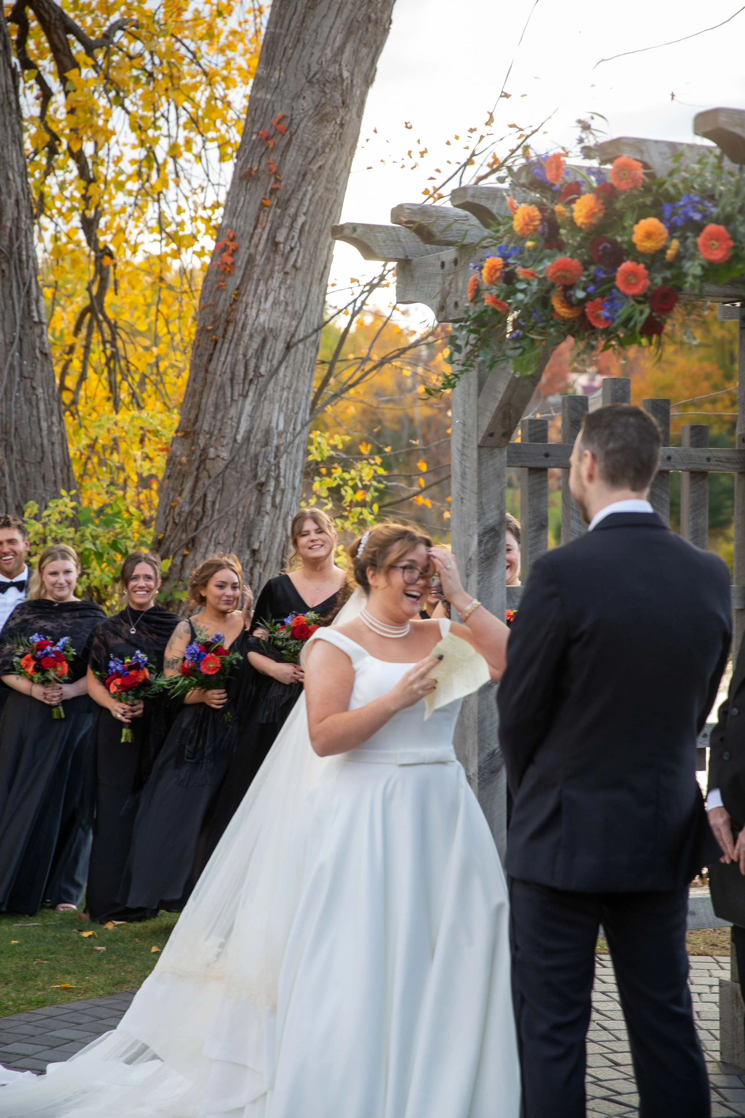 A bride and groom exchange vows during outdoor wedding ceremony in autumn, with bridesmaids and groomsmen in the background.