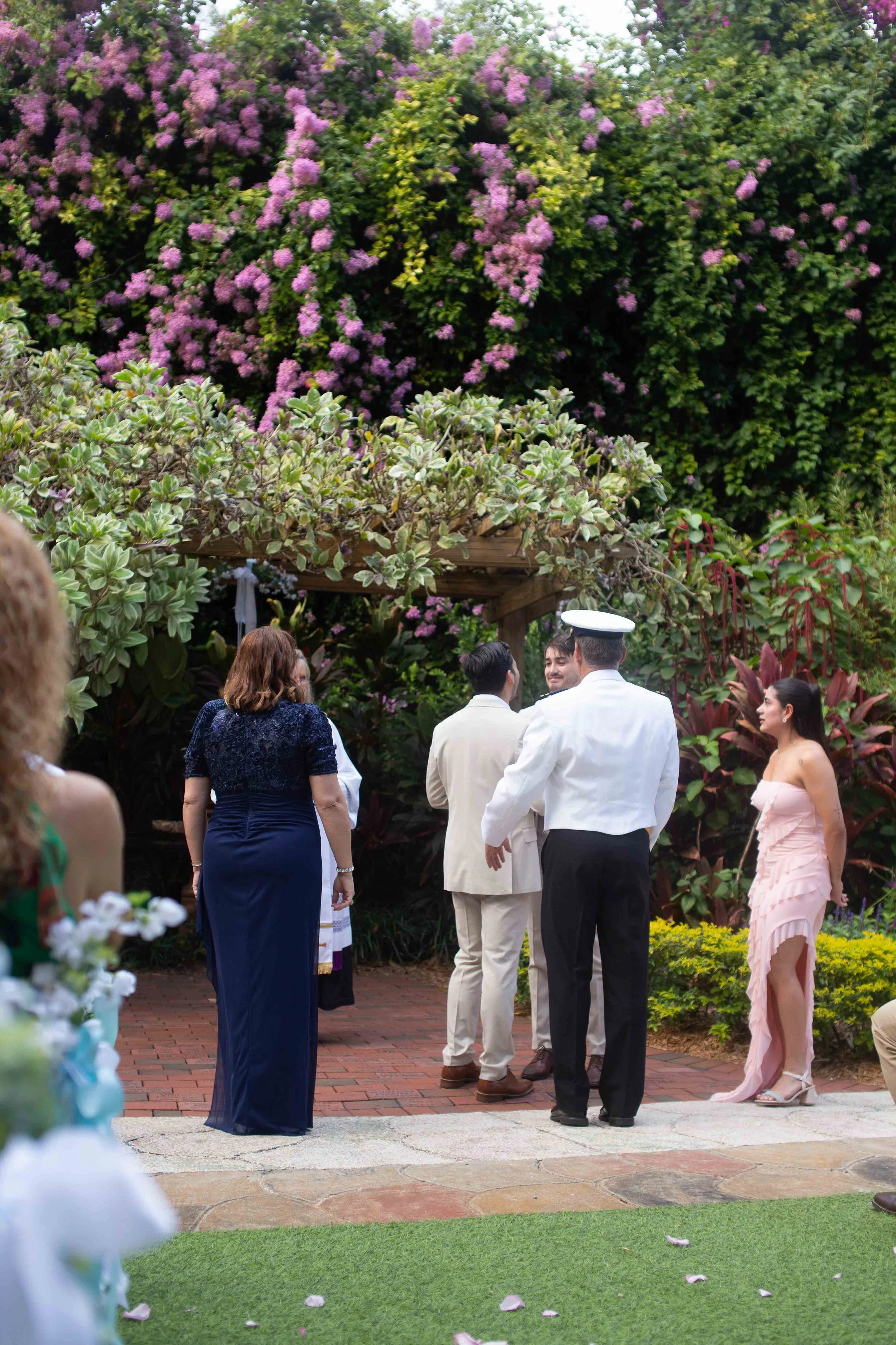 A wedding ceremony taking place outdoors in a garden with lush greenery and pink flowers. Several people, including a woman in a pink dress and a man in a white military uniform, stand around a couple at the altar.