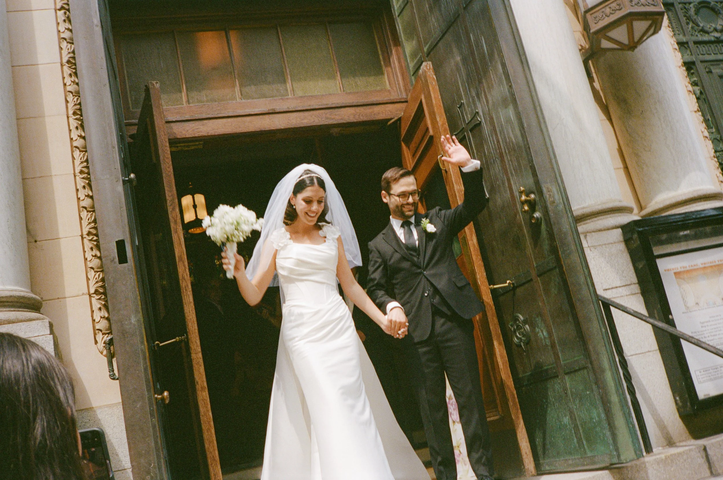 A bride and groom holding hands and smiling as they exit a church, with the groom waving, during their wedding ceremony.