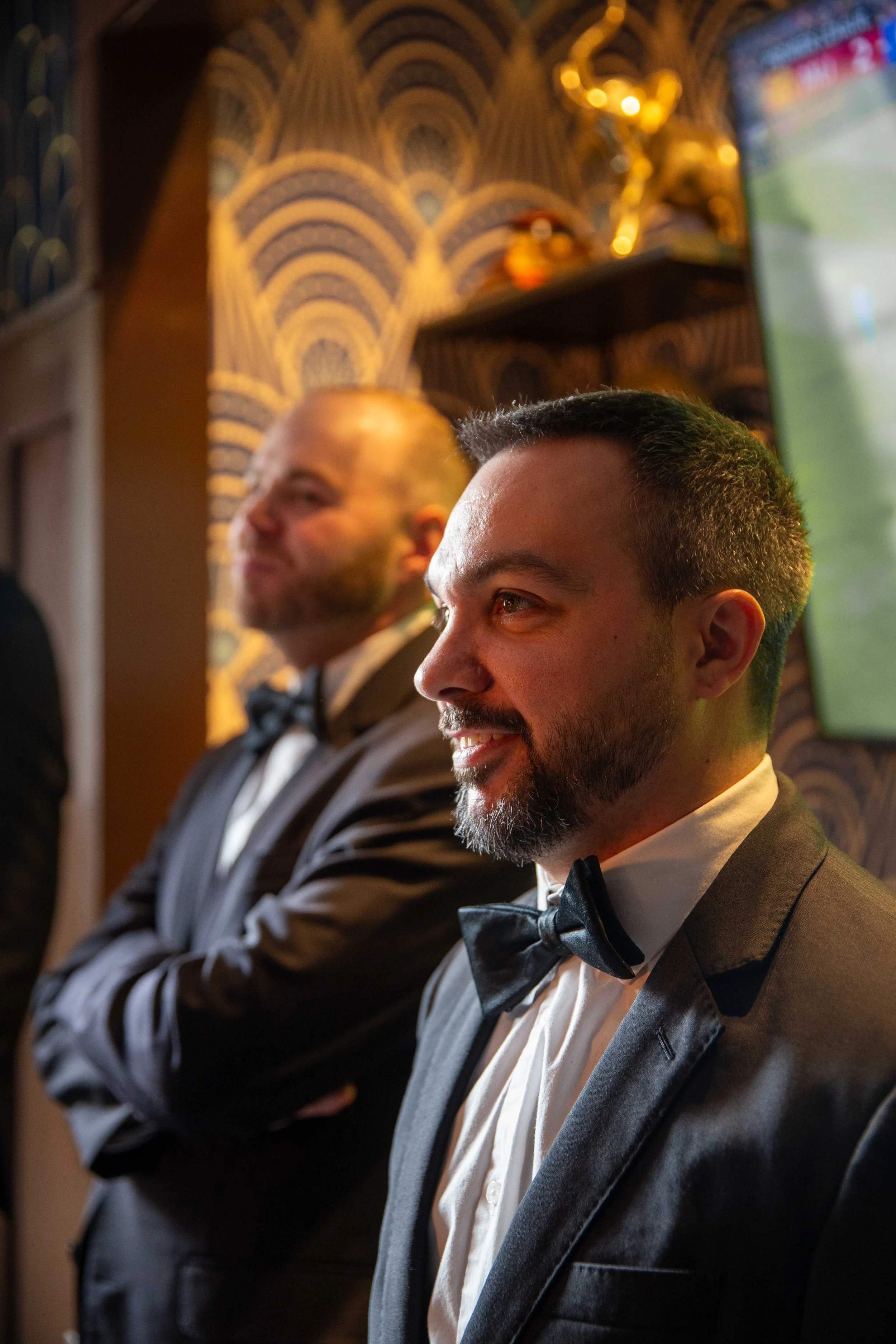 Two men in tuxedos with bow ties, standing indoors with patterned wallpaper and shelves with trophies, engaged in an event or celebration.