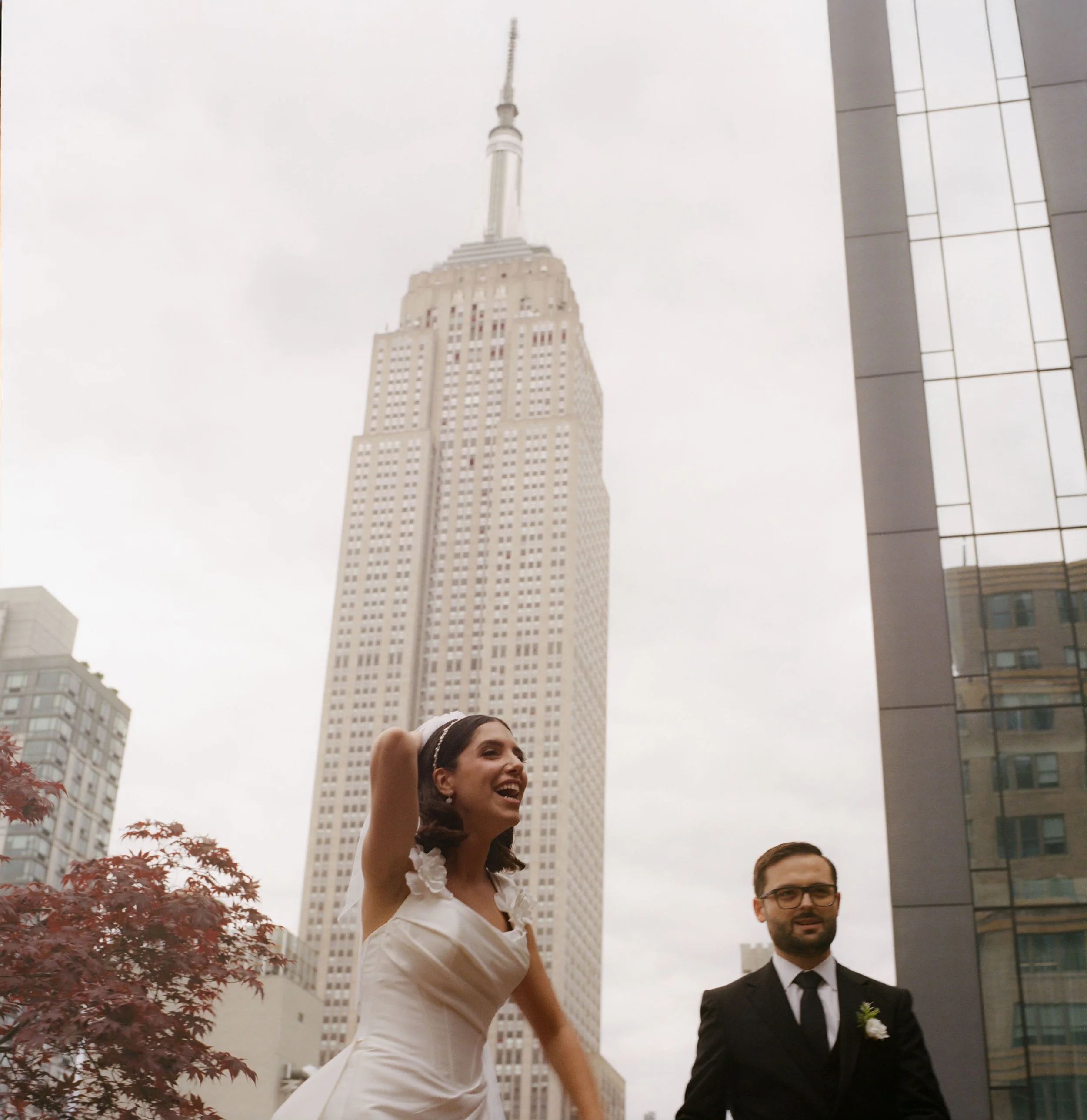 A woman in a white dress and a man in a suit are standing outside in front of the Empire State Building in New York City, with a cloudy sky overhead.