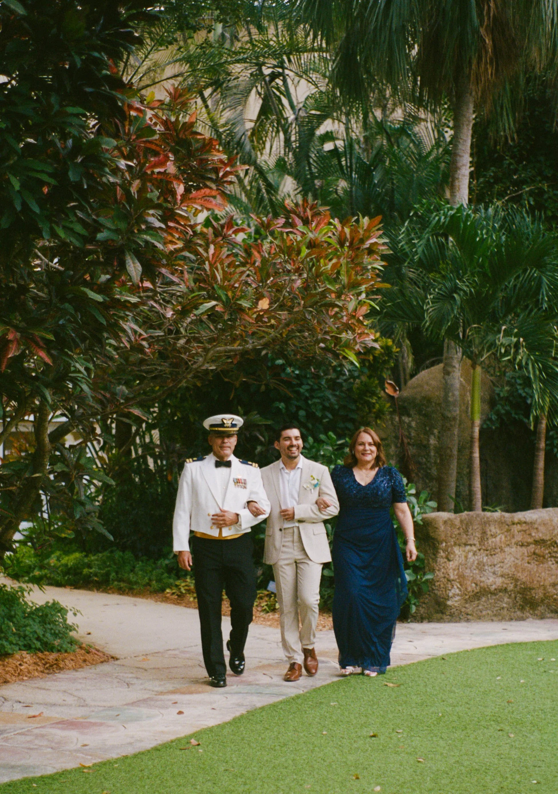 Three people walking arm-in-arm outdoors in a garden with lush green trees and plants, one in a military uniform, one in a light-colored suit, and one in a dark blue dress.
