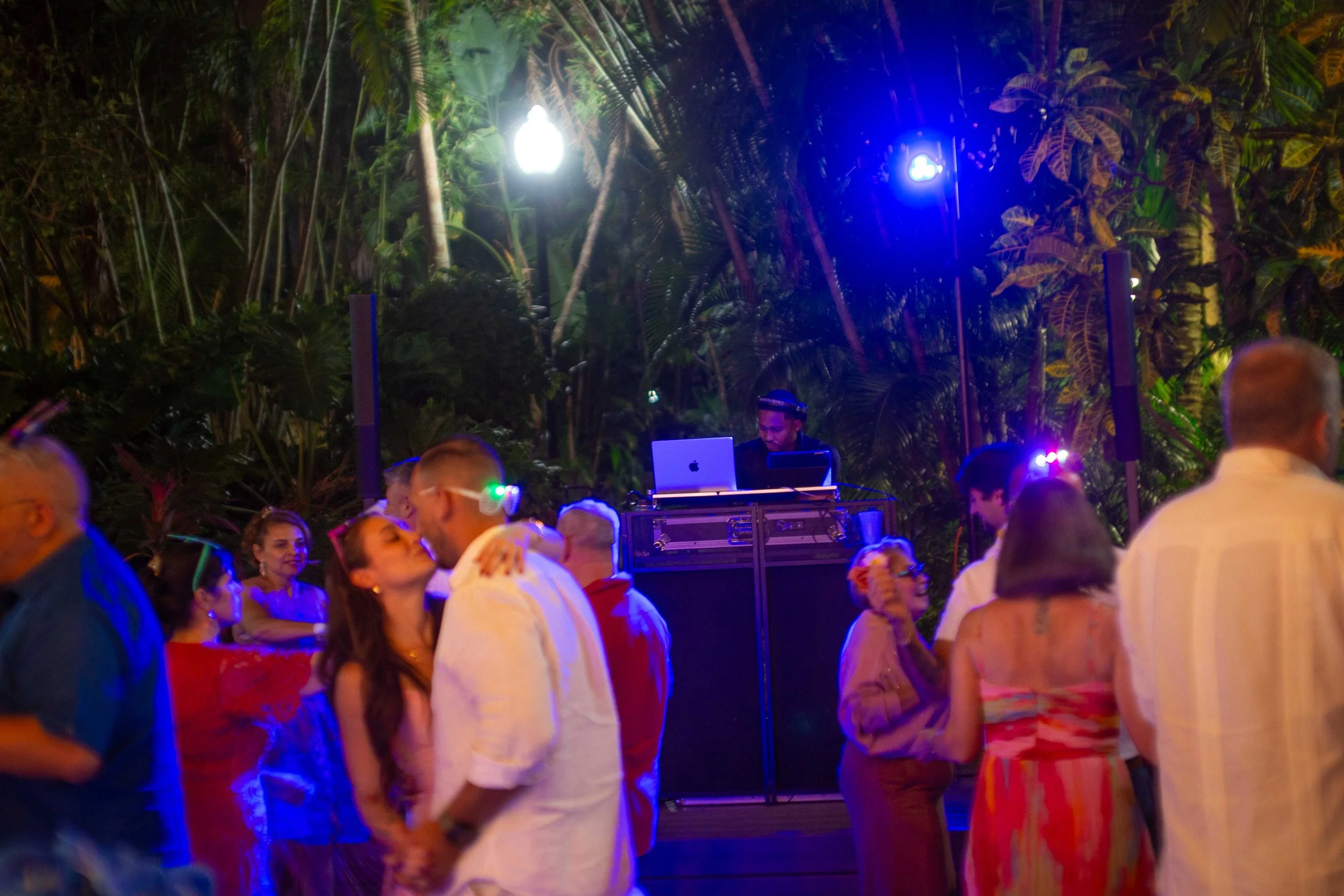 People dancing and socializing at an outdoor party or event with a DJ playing music, surrounded by tropical plants and illuminated by colorful lighting.