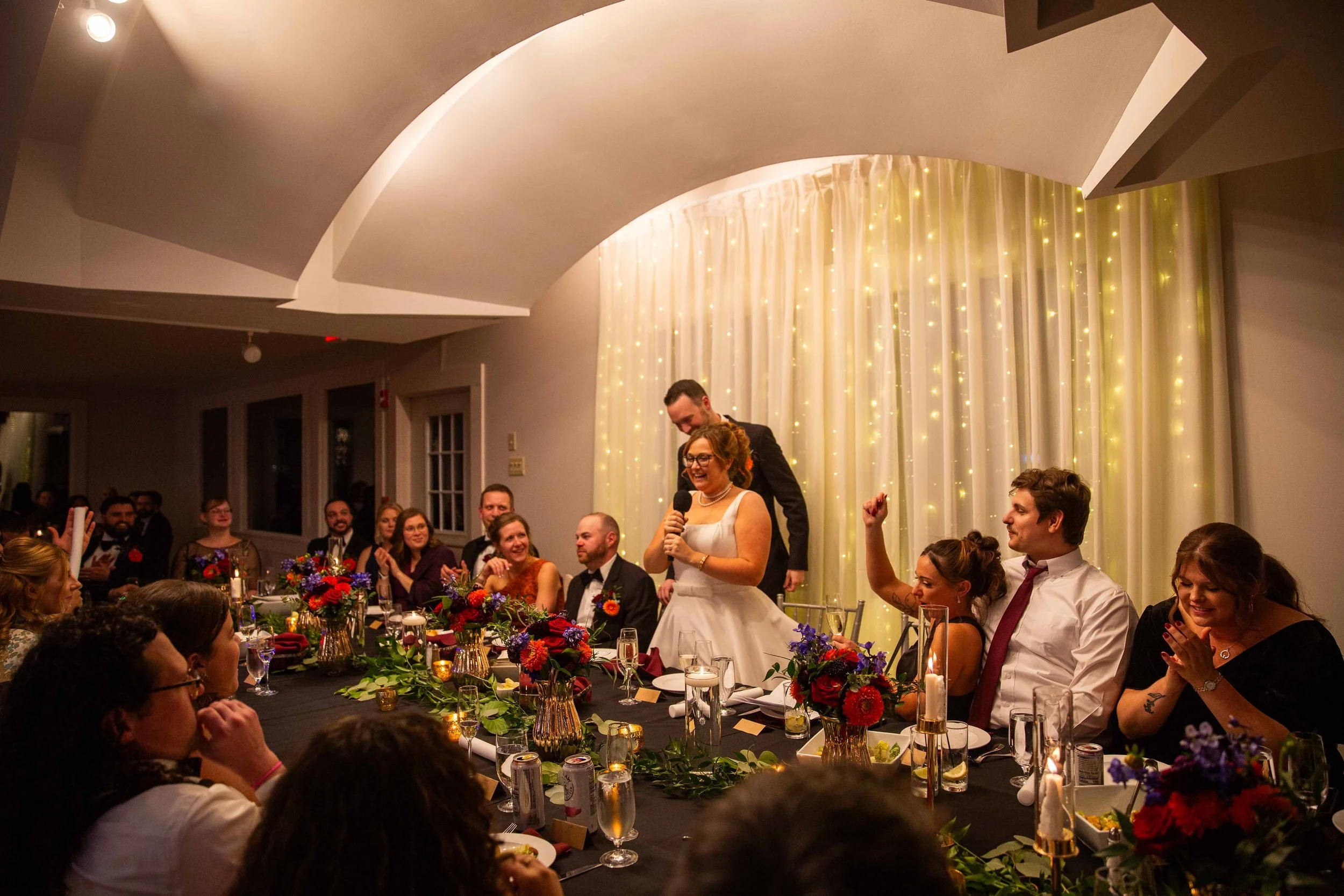 A wedding reception with guests sitting at a decorated table, a bride speaking into a microphone, and a groom standing behind her, in a room with string lights and white curtains.