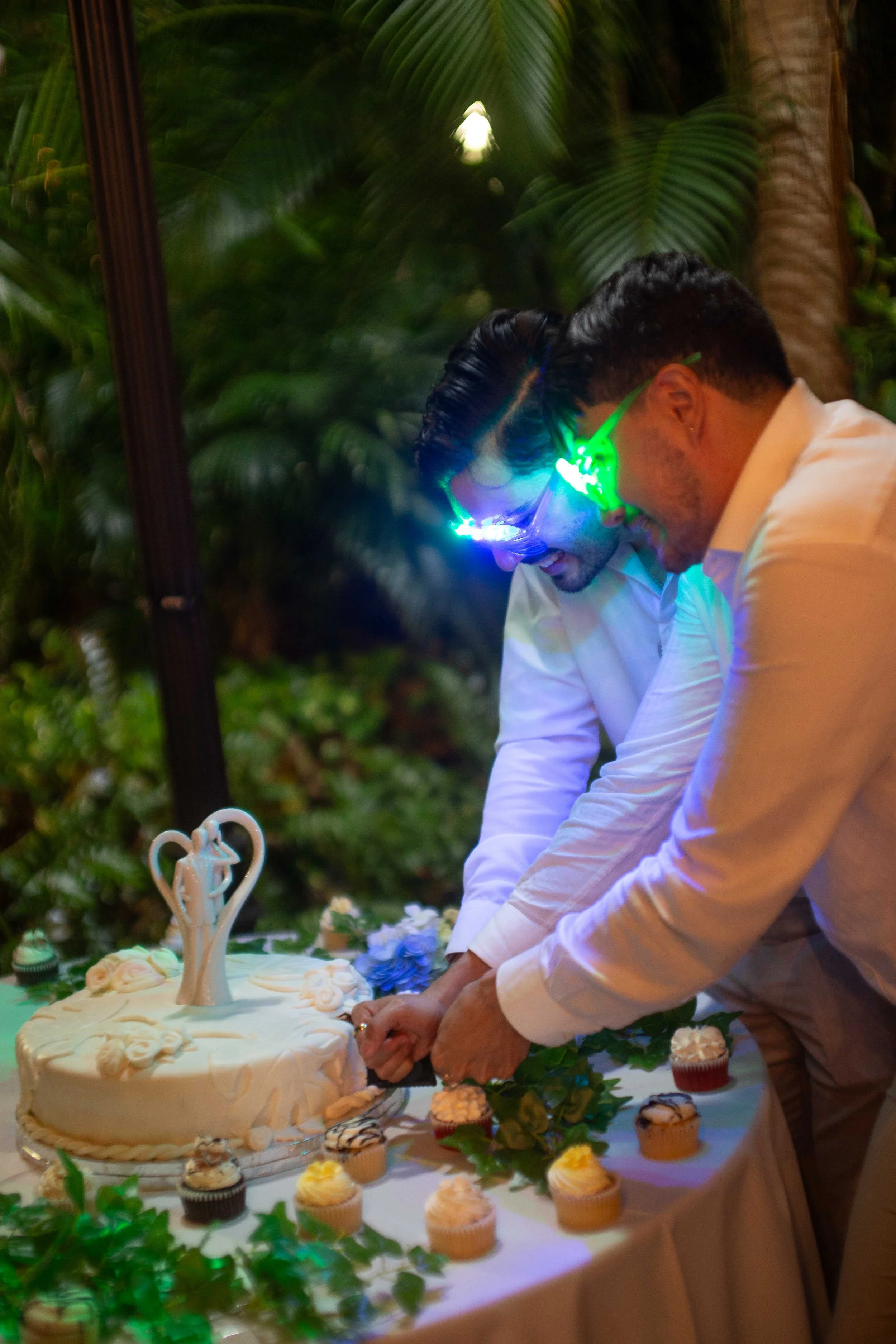 Two men cut a wedding cake together at a celebration, wearing glow-in-the-dark glasses, surrounded by cupcakes and greenery.