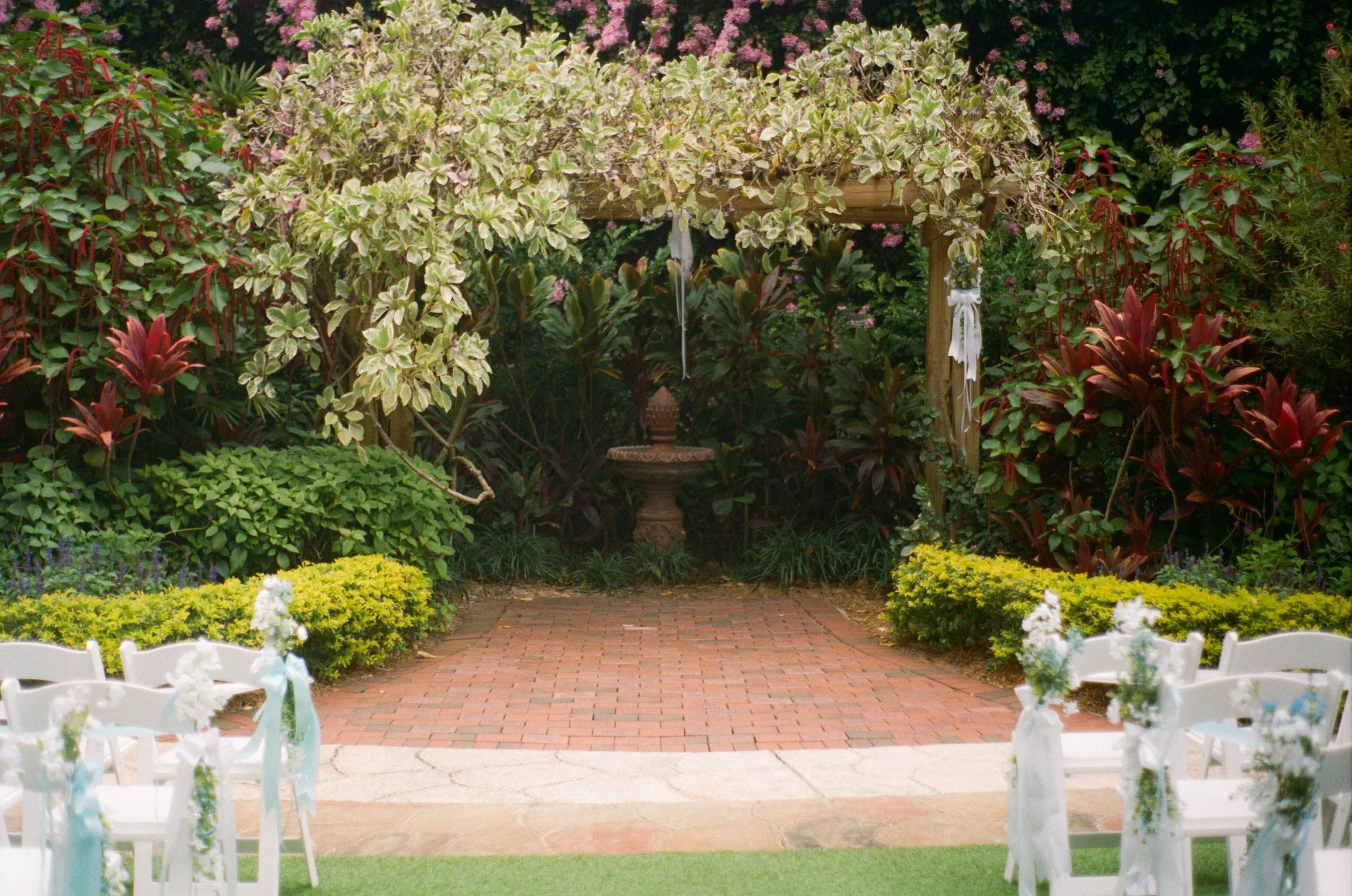 A small outdoor wedding ceremony setup in a lush garden with white chairs decorated with flowers and ribbons, facing an archway adorned with greenery and flowers, with a brick pathway leading to a decorative fountain in the background.
