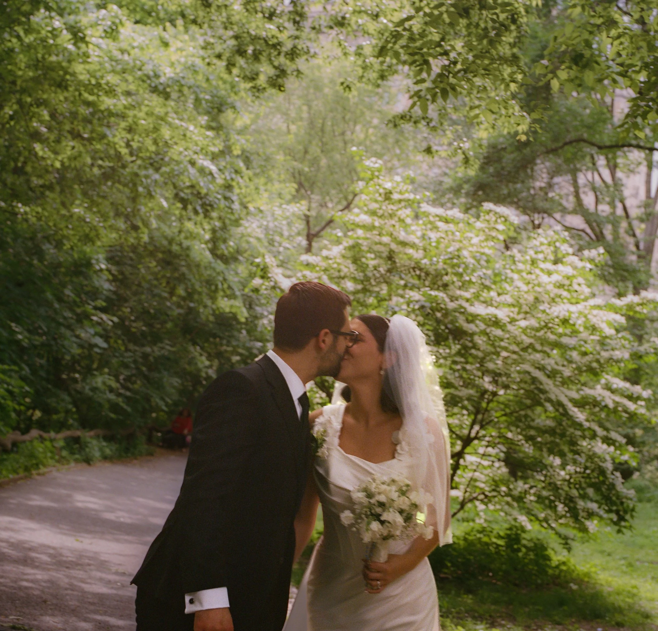 A bride and groom kissing outdoors in a green park, surrounded by blossoming trees, on their wedding day.