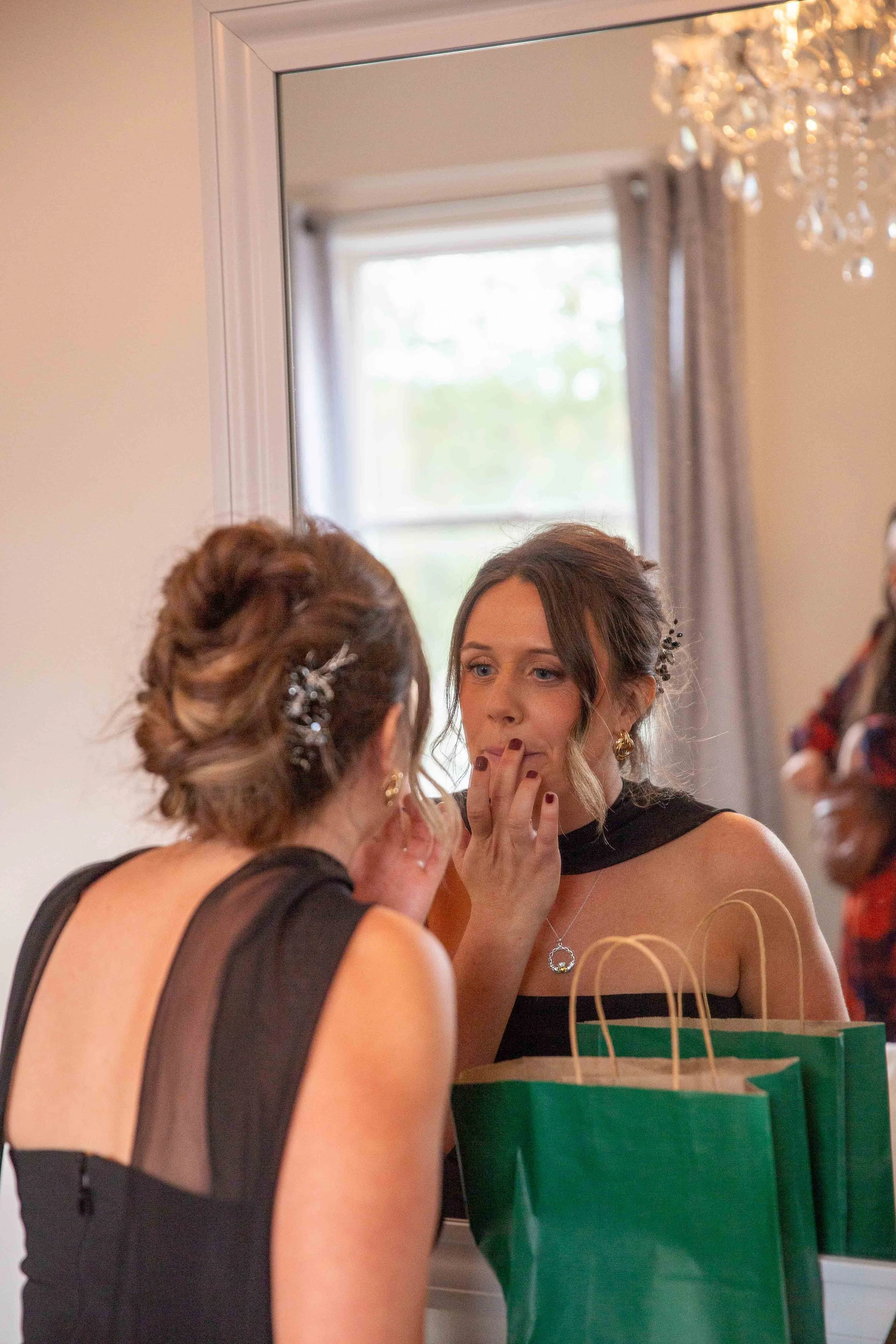 A woman looking into a mirror, adjusting her lipstick while preparing for an event, with gift bags on the table in front of her.