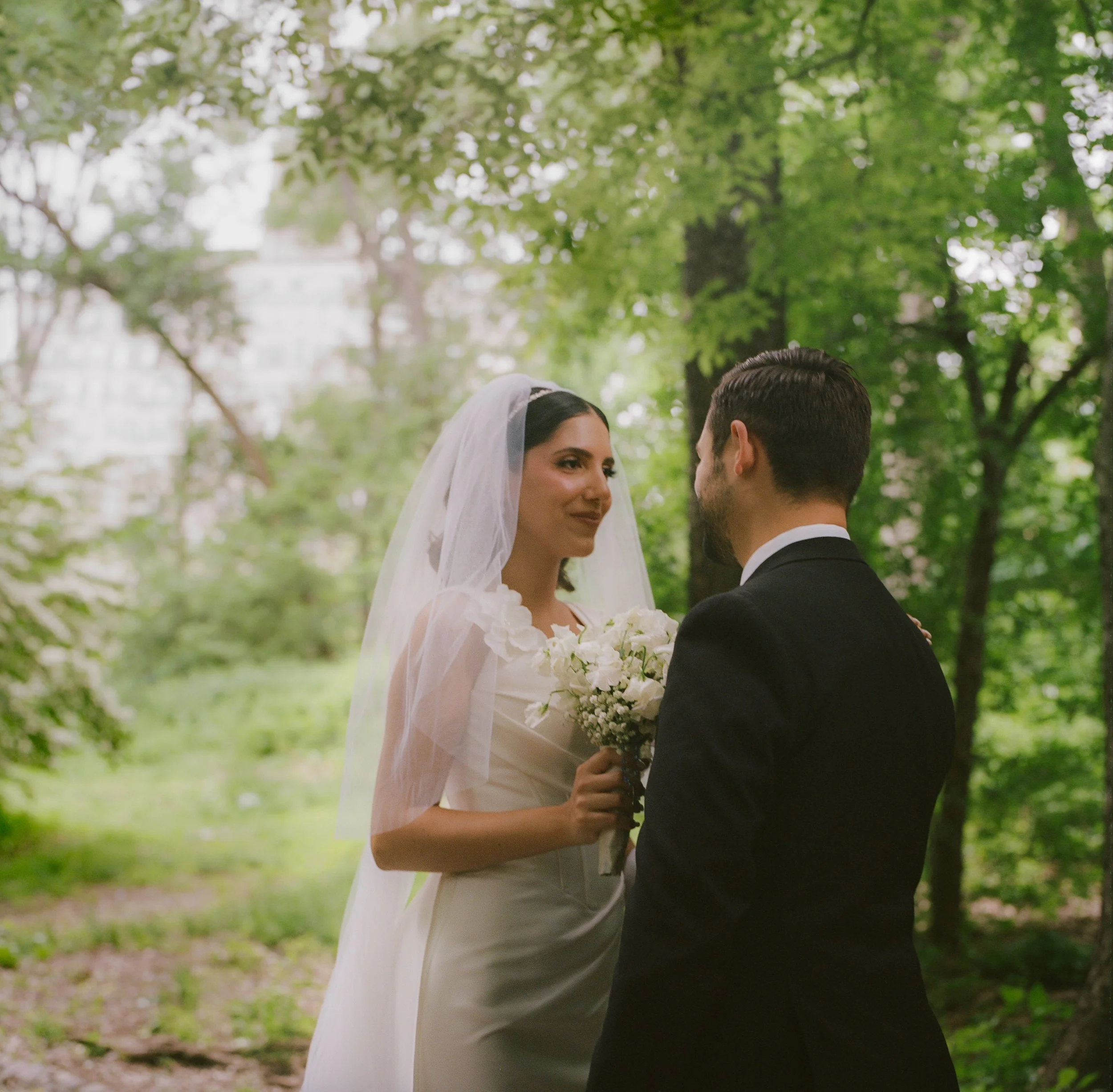 A bride and groom stand face to face in a lush green outdoor setting with tall trees. The bride wears a white wedding dress and veil, holding a bouquet of white flowers. The groom wears a black suit. They gaze into each other's eyes with a blurred ci