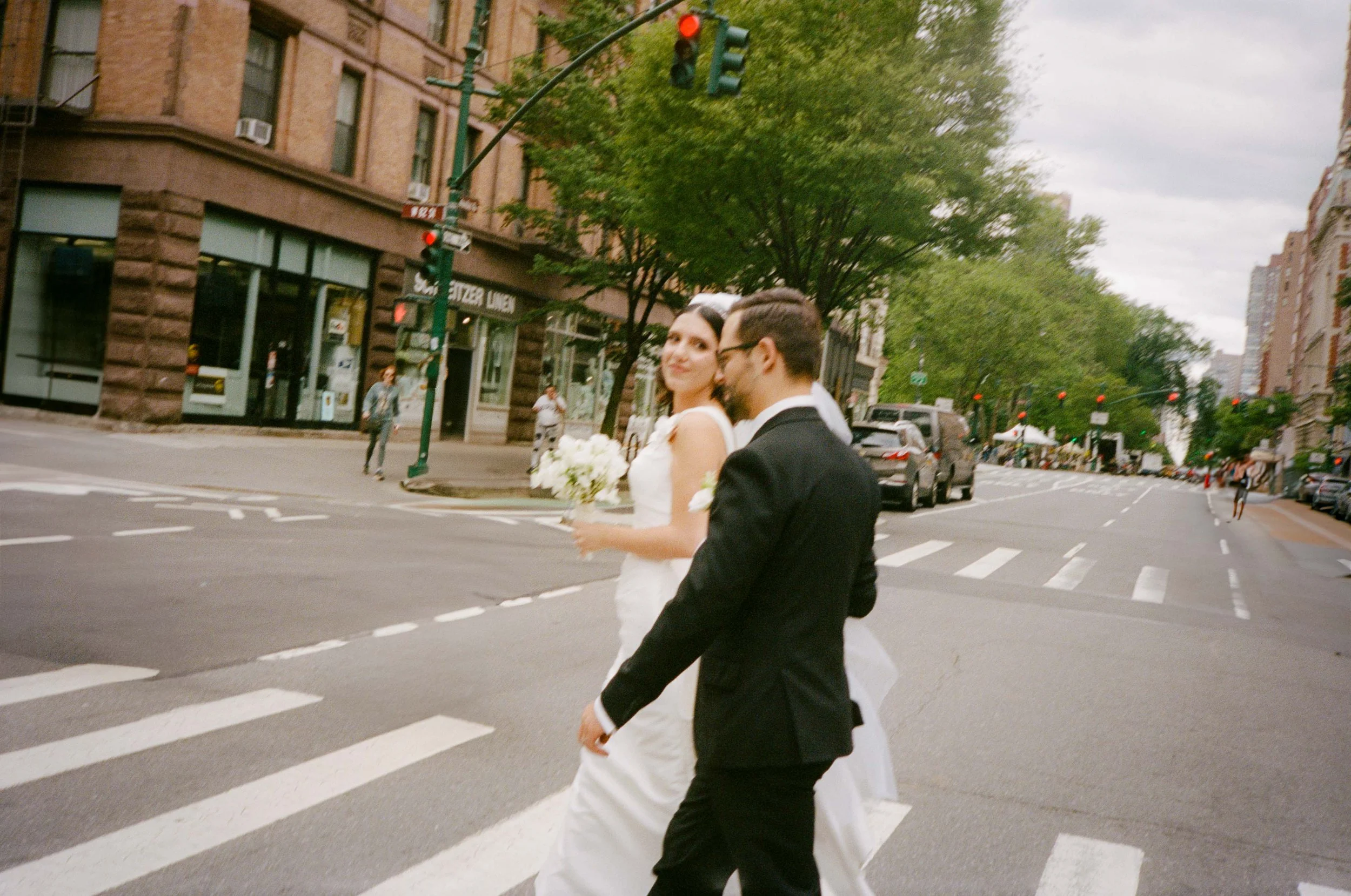 Bride in white wedding dress holding a bouquet, walking with groom in black tuxedo across a city street at crosswalk, surrounded by buildings and trees.