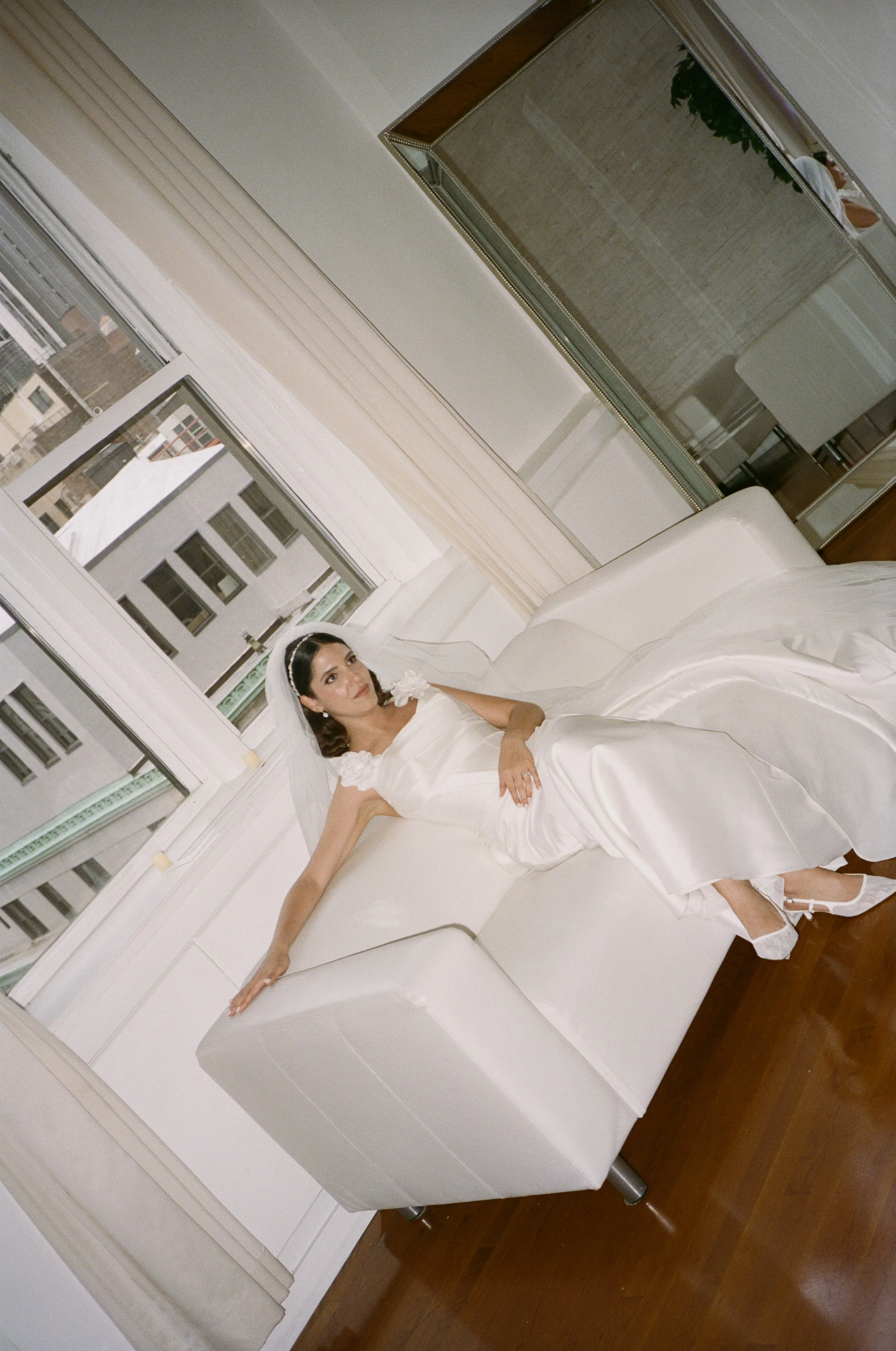 A woman in a white wedding dress sitting on a white sofa in a bright room with large windows.